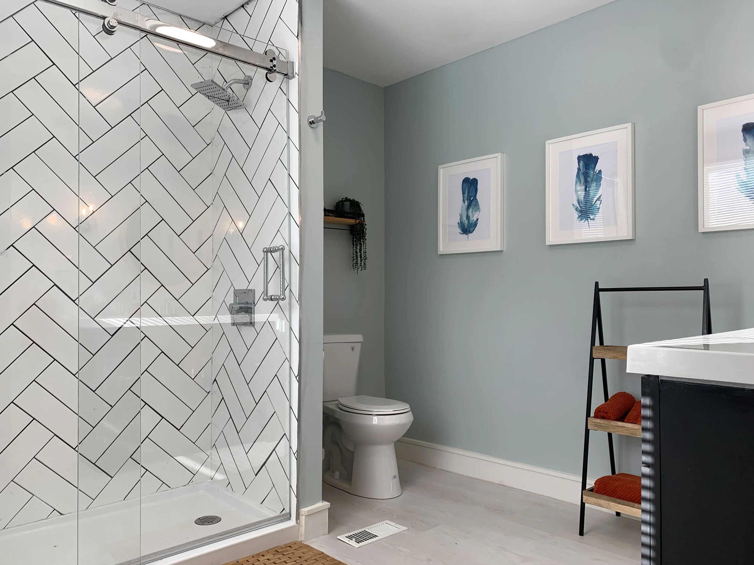 A bathroom with a glass-enclosed shower featuring white tiles arranged in a herringbone pattern and a rainfall showerhead. To the right, there's a toilet, framed artwork of blue feathers on a light blue wall, a black chair with wooden shelves holding