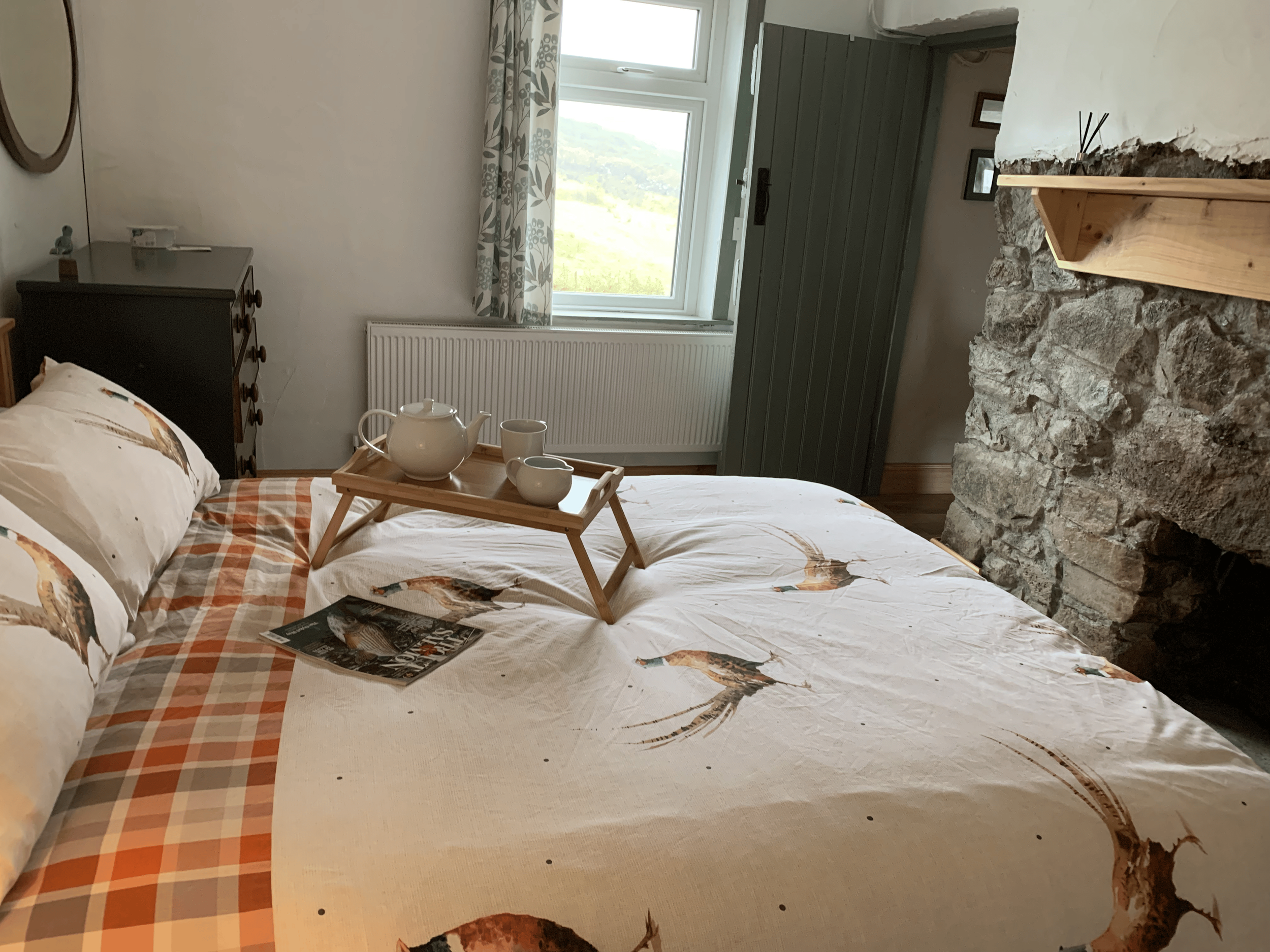 A cozy bedroom with a bed covered in a furniture-themed duvet and matching pillowcases, topped with a wooden breakfast tray holding a teapot and cups, near a stone fireplace with a wooden mantle, a window with floral curtains showing a scenic landsca