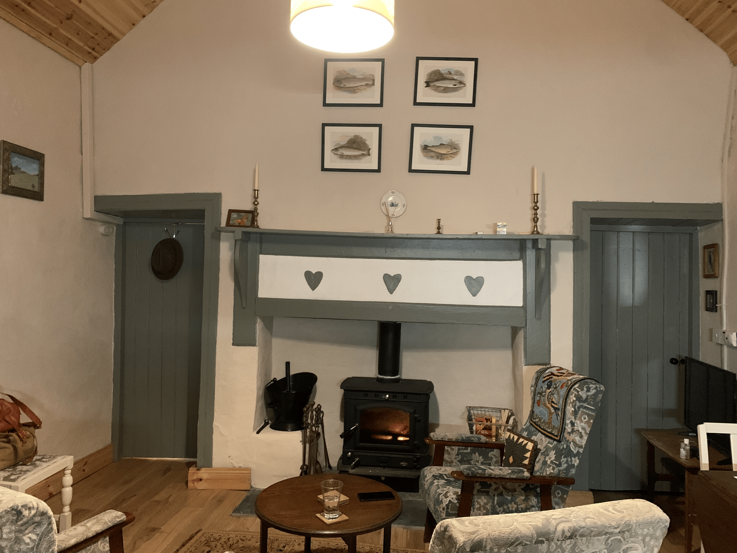 A cozy living room with a wood stove, a sofa with patterned upholstery, and framed artwork of boats on the wall above the fireplace, under a wooden ceiling with a ceiling light.