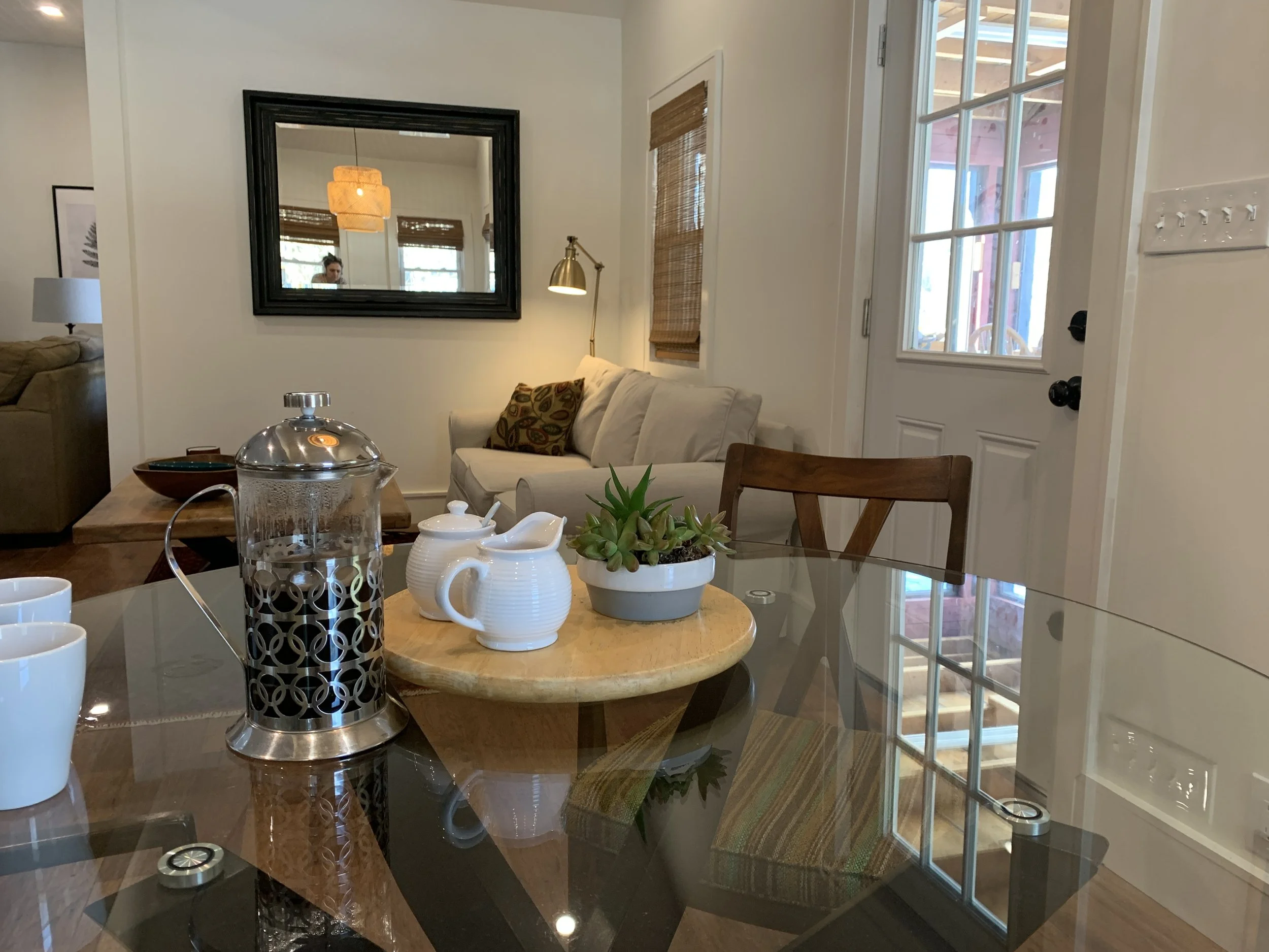 Interior view of a living room and dining area with a round glass-top table, a white couch with patterned pillows, a wall mirror, a brass desk lamp, and a door with glass panels leading outside. Decorations include a succulent plant on the table and 