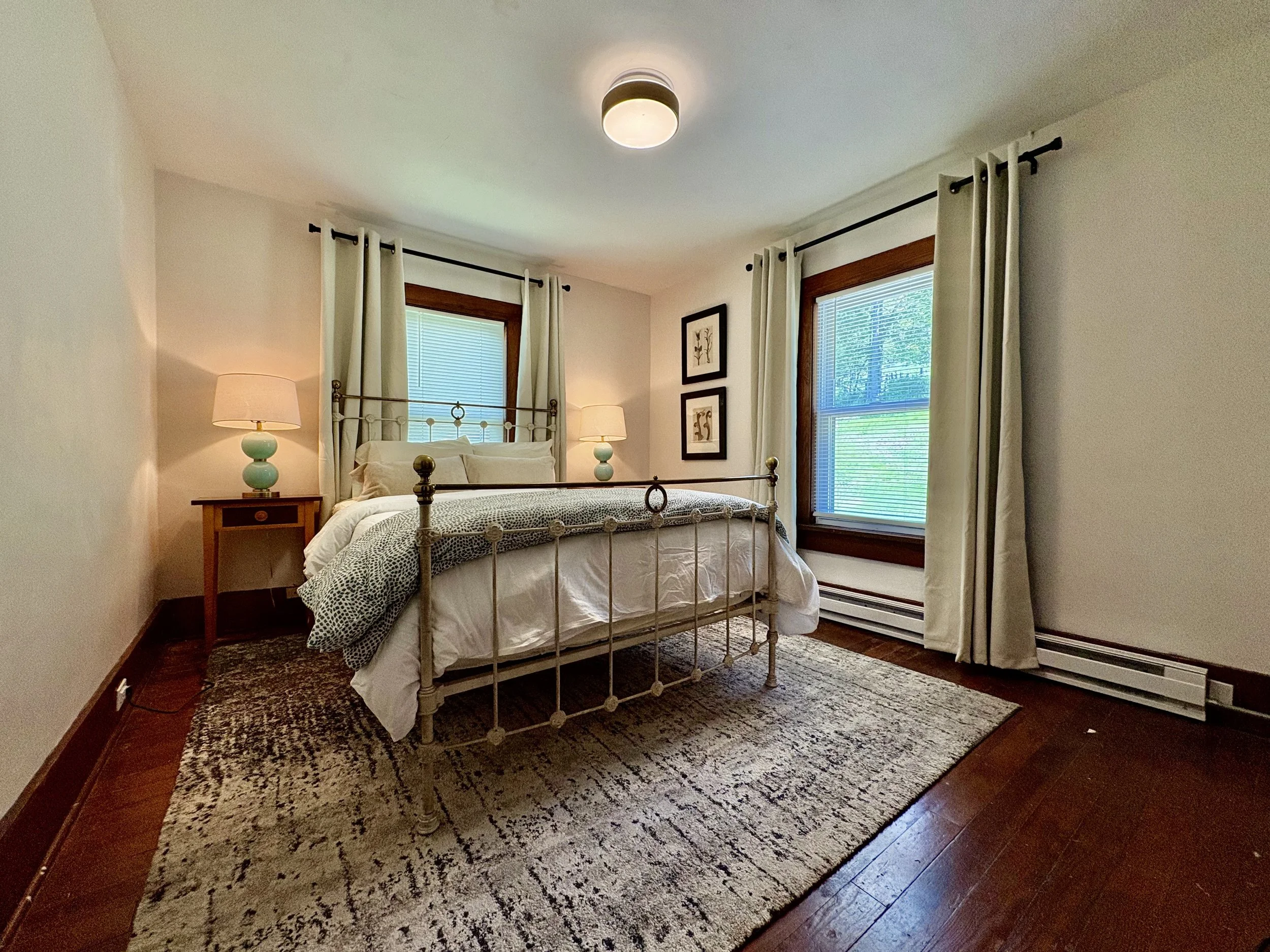 A bedroom with a metal bed frame, white bedding, two matching nightstands with turquoise lamps, two windows with white curtains and blinds, framed artwork on the wall, hardwood floor, and an area rug.