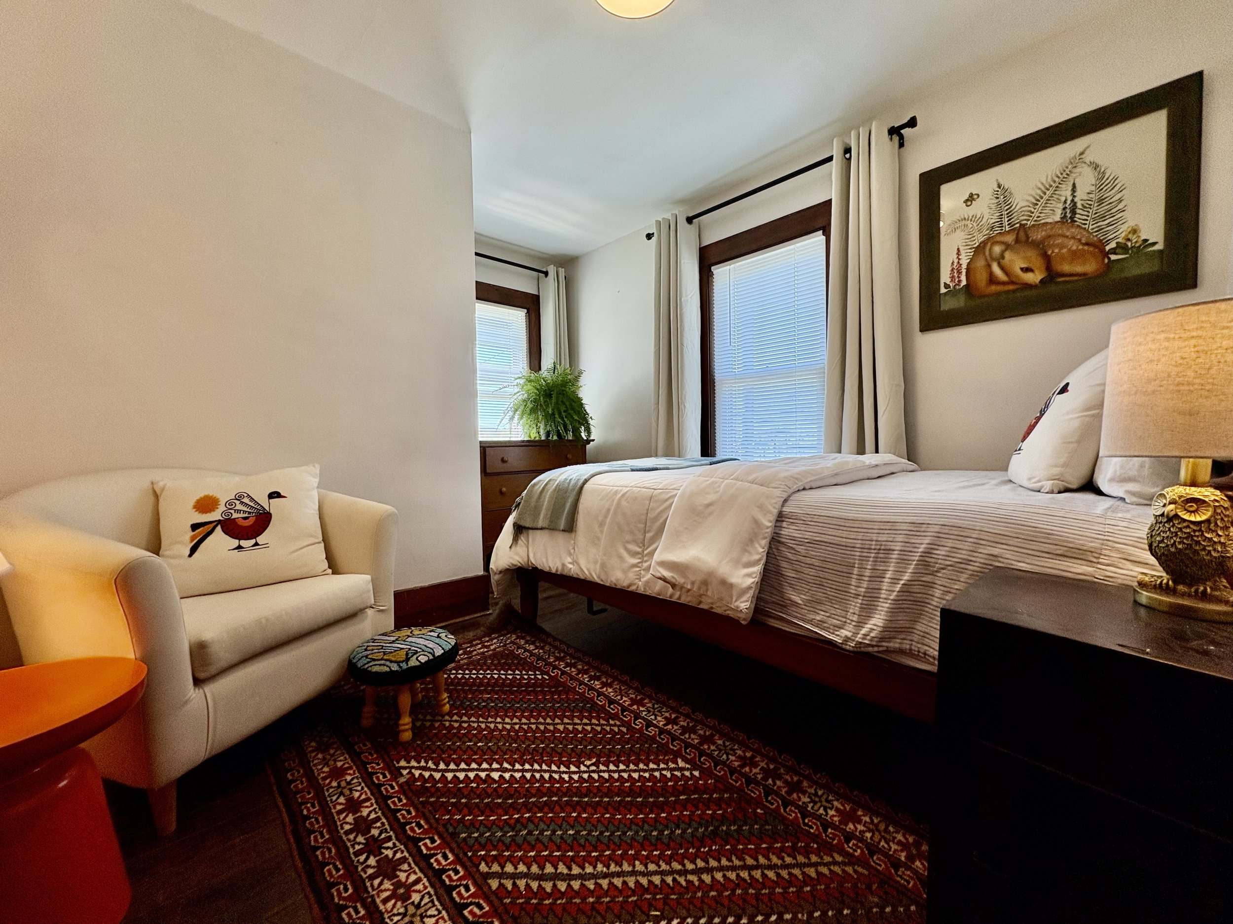 A cozy bedroom with a bed, framed picture of a sleeping animal on the wall, large windows with white curtains, and a small sofa with a decorative pillow featuring a bird design. There is a colorful patterned rug on the dark wooden floor, a potted fer