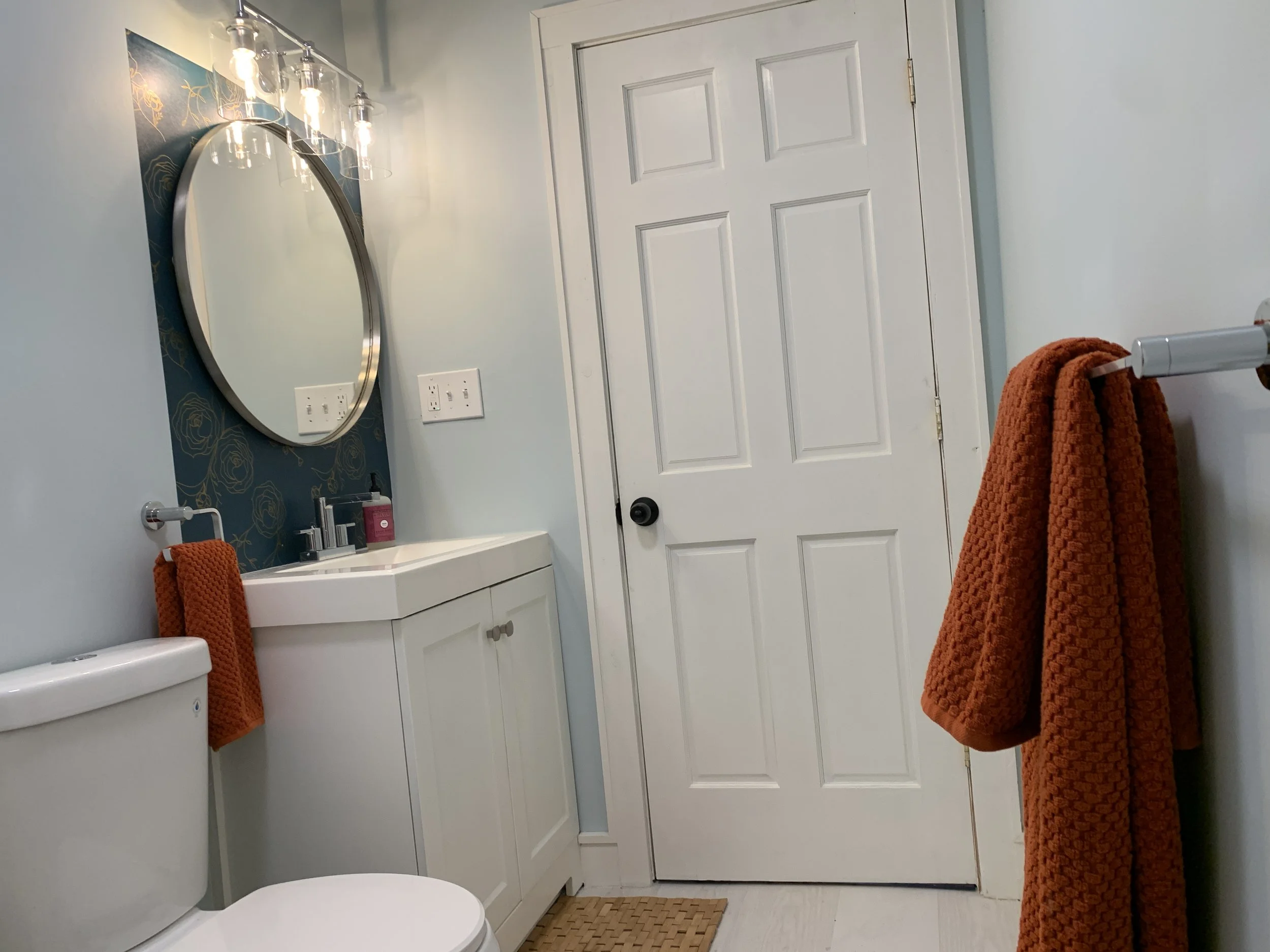 A bathroom with a white door, a small white sink with a mirror above it, orange towels hanging on a towel bar, a white toilet, and a mat on the floor. The wall above the sink has blue wallpaper with gold floral patterns and light fixtures with glass 