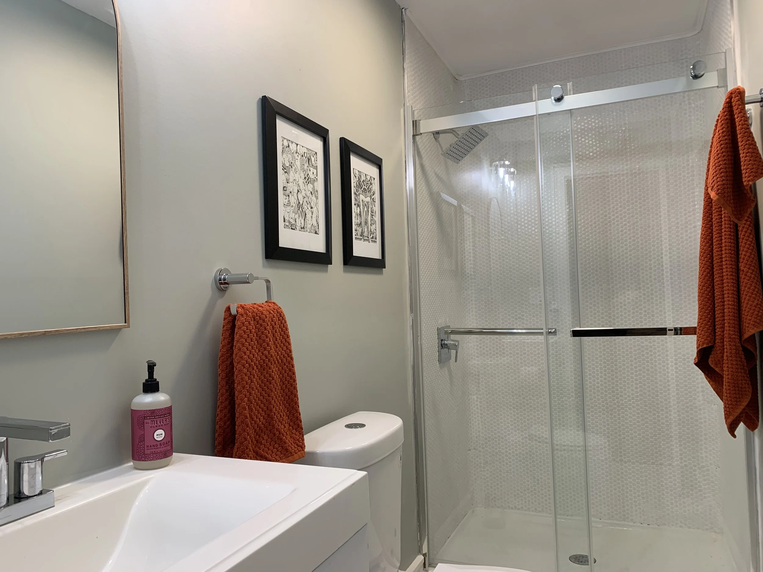 A bathroom featuring a wall-mounted mirror, framed black-and-white artwork, orange towels, a white vanity with a soap dispenser, a white toilet, and a glass-enclosed shower with a patterned wall and a rain showerhead.