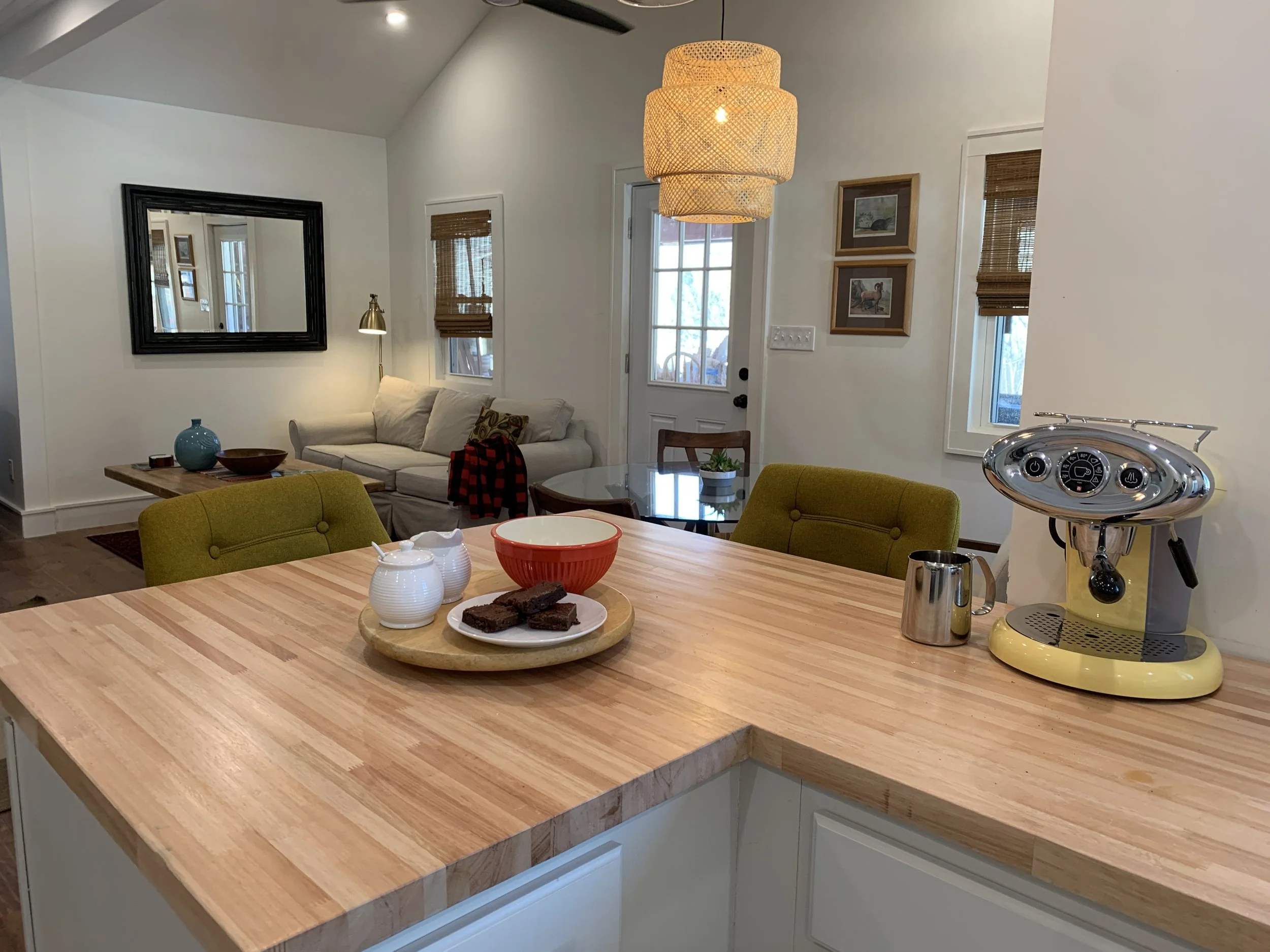 A view from a kitchen island looking into a living room with a beige sofa, a black framed mirror, and windows with bamboo blinds. There is a wooden table with a potted plant, a bowl of brownies, and a yellow espresso machine.