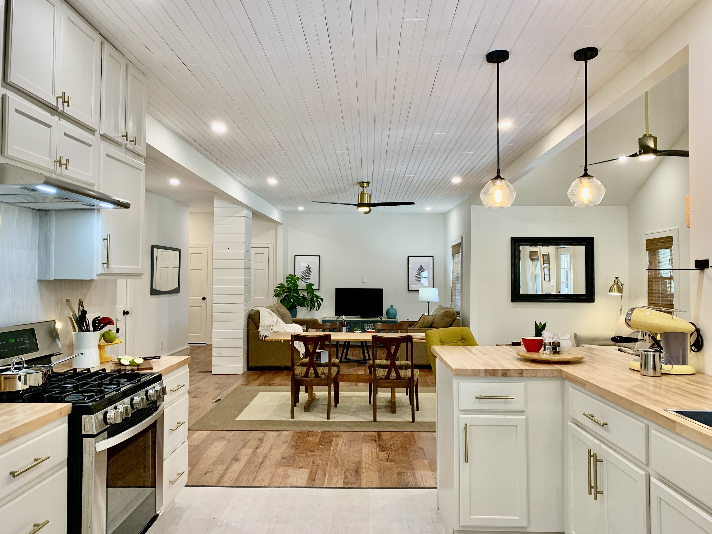 Interior view of a modern kitchen opening into a living room with white cabinetry, a gas stove, kitchen island, dining table, and living room furniture including sofas and a lamp, with pendant lights and a ceiling fan.
