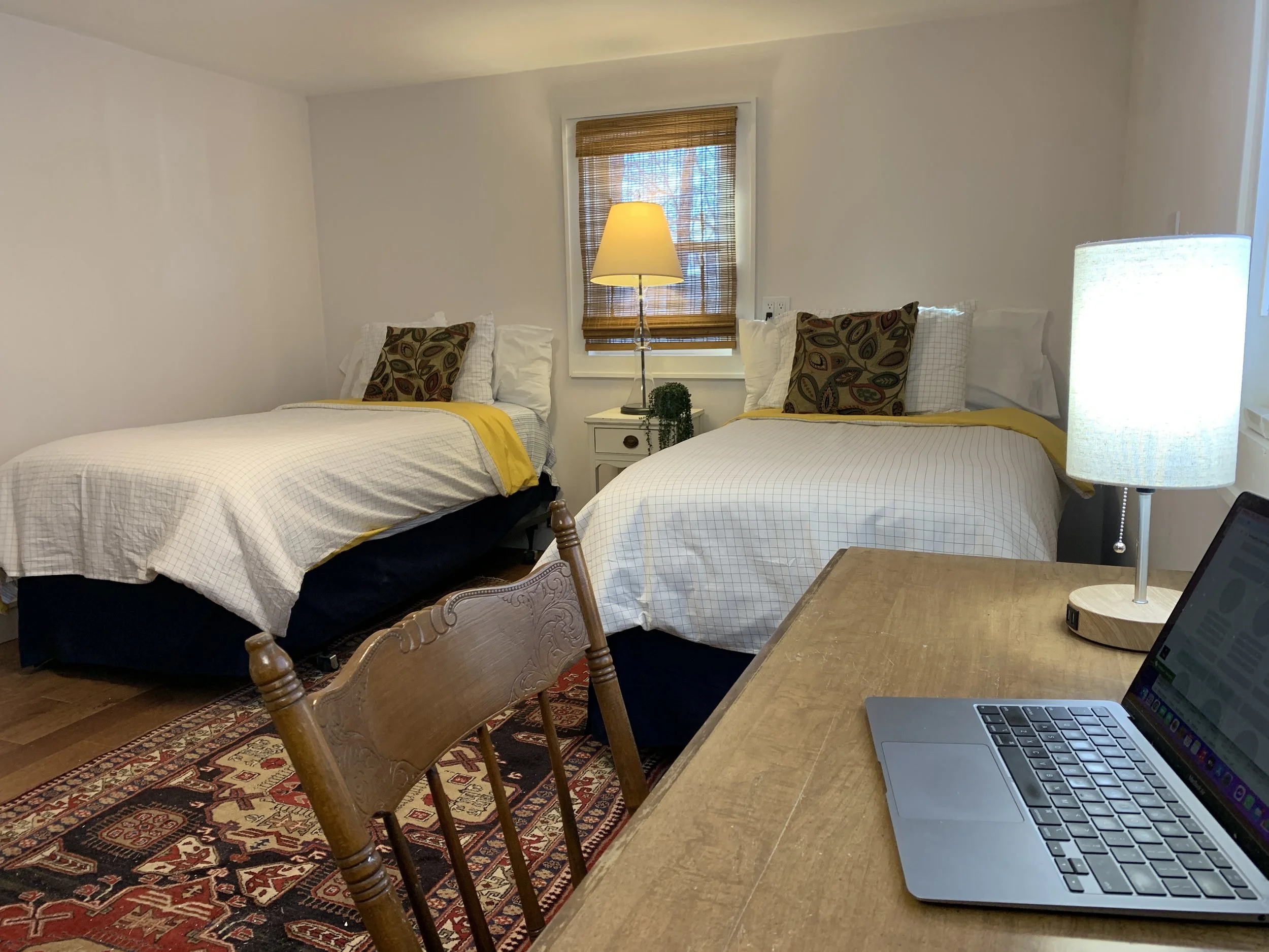 A bedroom with two twin beds with white checkered bedding and decorative pillows, a nightstand with a lamp and plant, a window with bamboo blinds, a lamp, and a laptop on a wooden table, with a patterned rug on the hardwood floor.