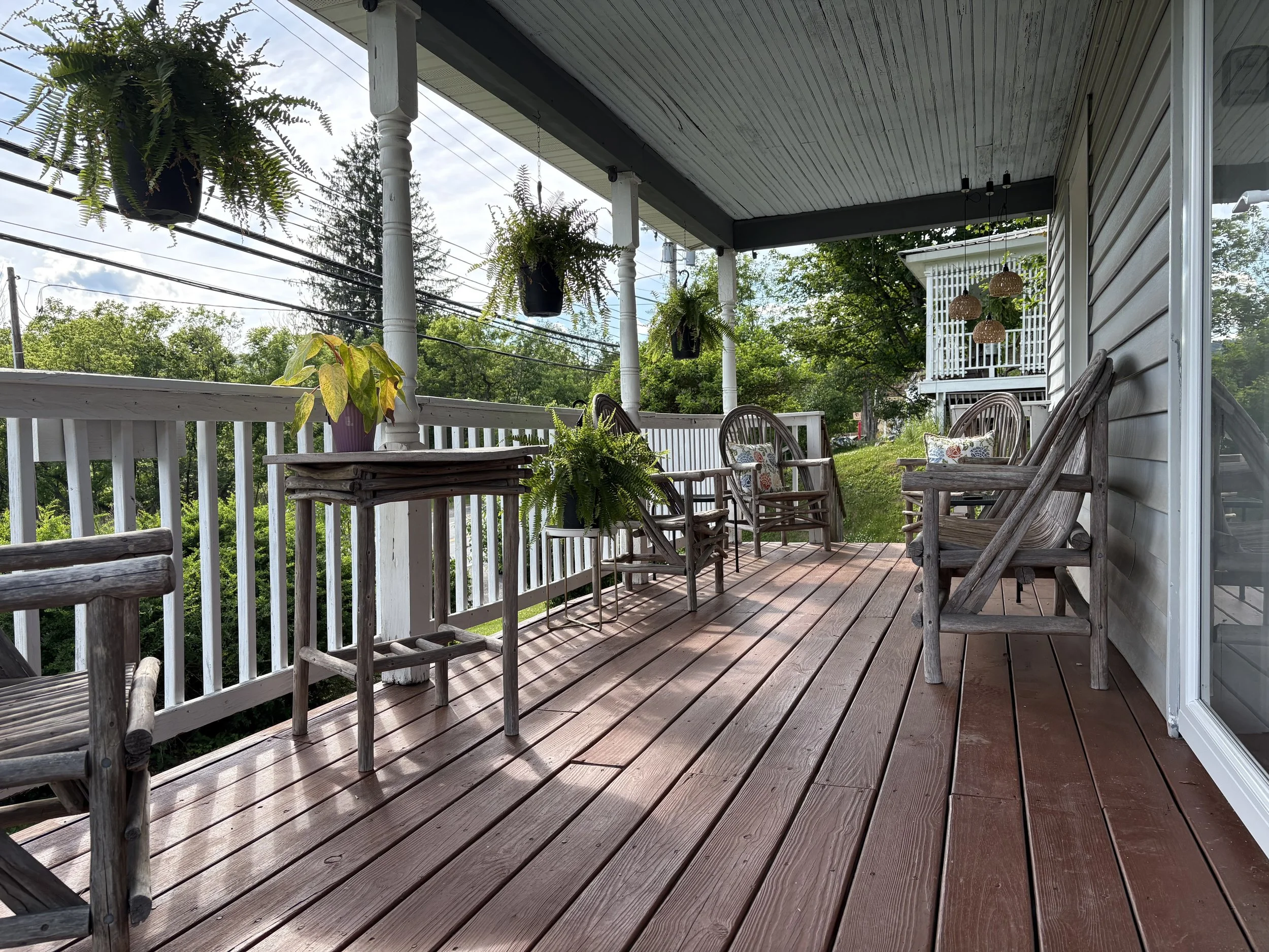 A wooden porch with white porch railing, several hanging potted ferns, and outdoor seating including wooden chairs with floral cushions and a wooden table with a potted plant, surrounded by green trees and a neighboring house with a balcony in the ba
