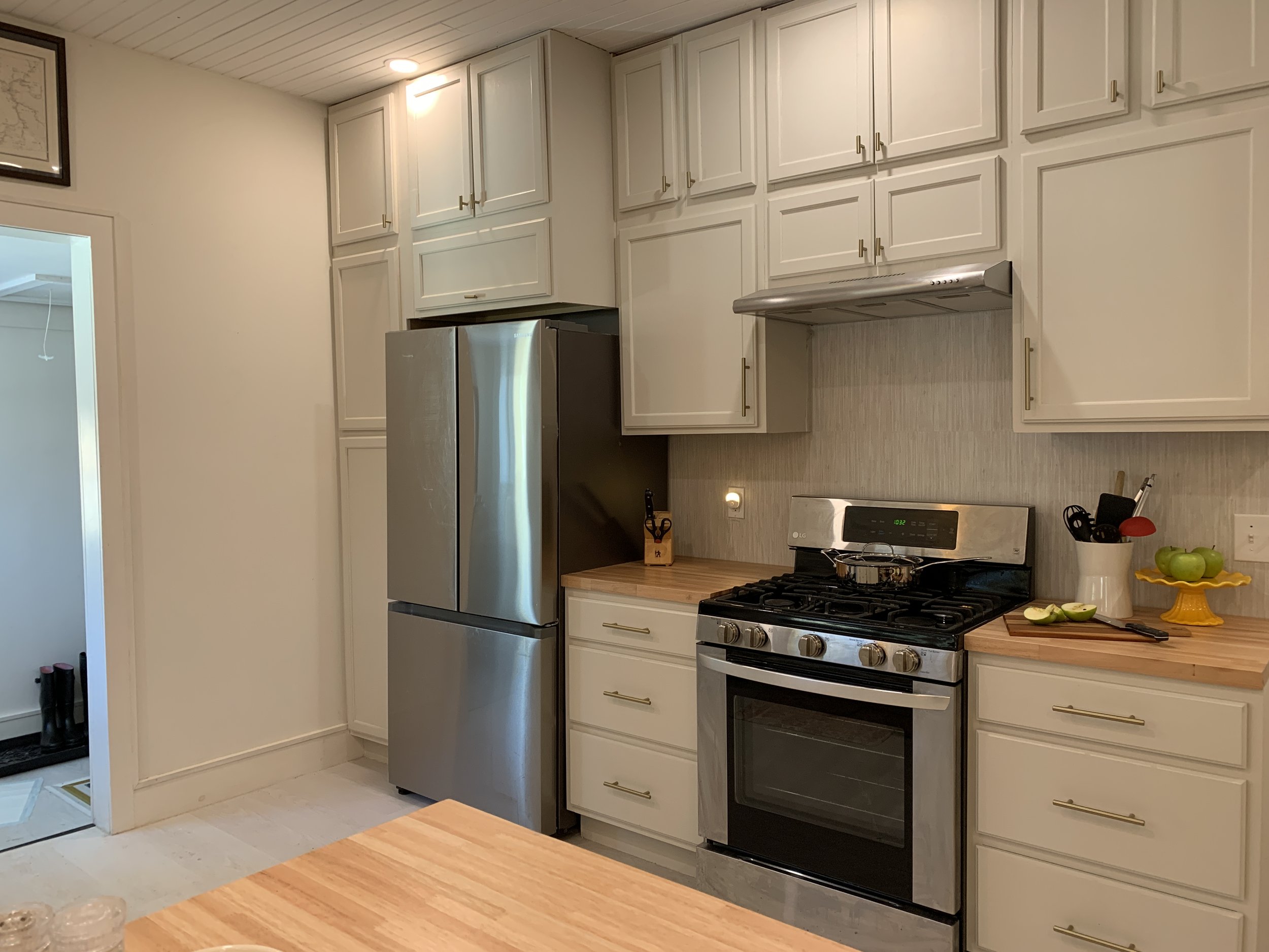 A modern kitchen with white cabinets, a stainless steel refrigerator, a gas stove, and a light wood countertop with fresh green apples and a yellow cake stand. There's a picture frame on the wall and a doorway leading to another room.
