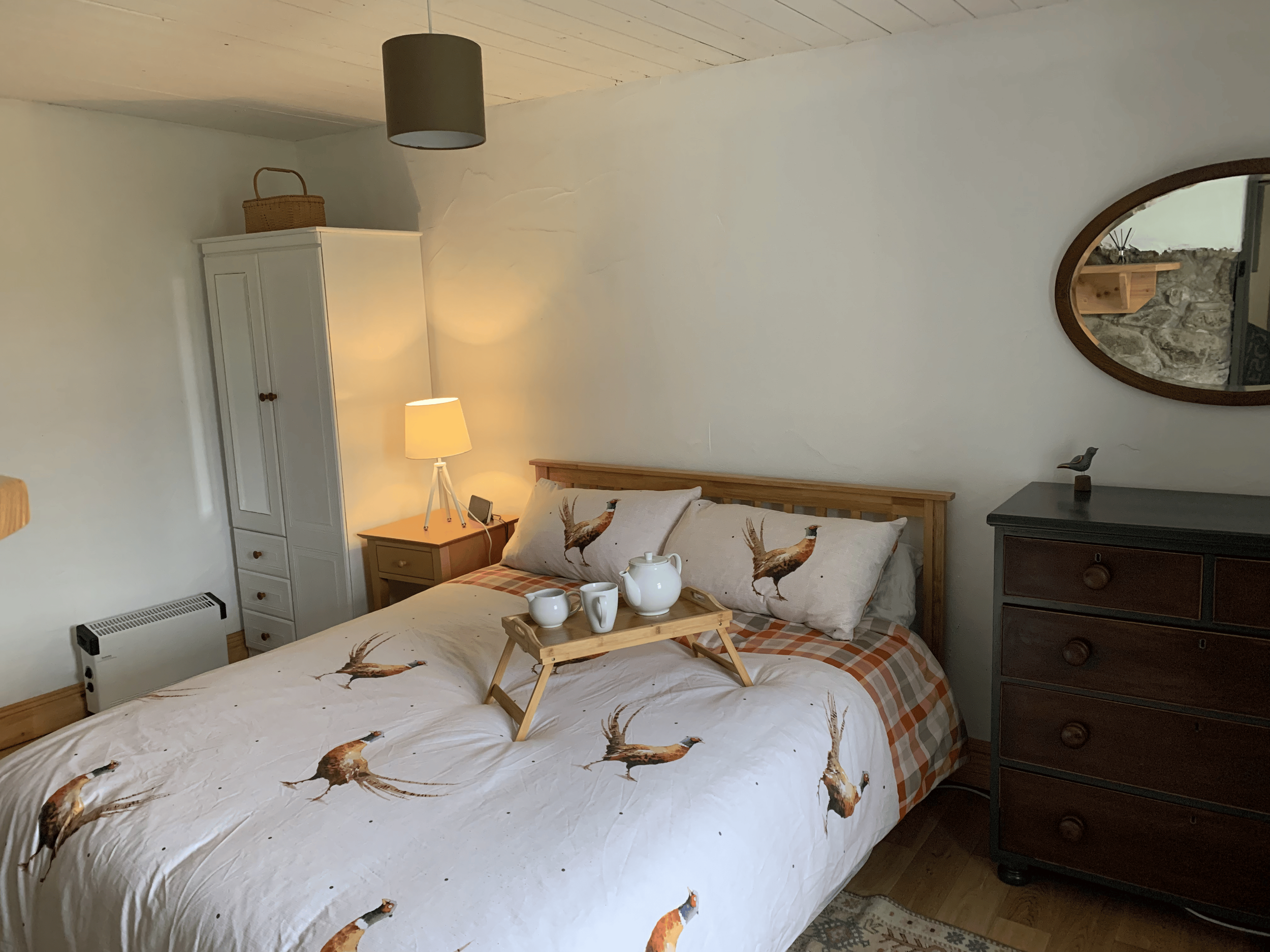 A cozy bedroom with a bed covered in a deer-patterned quilt, a wooden headboard, and matching pillows. Beside the bed is a small bedside table holding a tablet and a trip of white ceramic teapots. There is a black dresser with a decorative bird figur