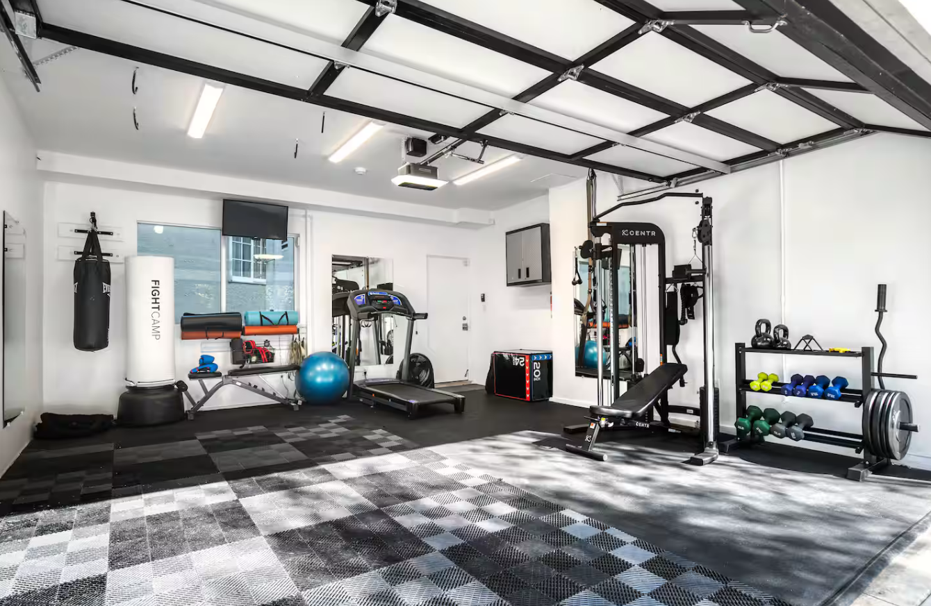 Home gym with various workout equipment including a punching bag, treadmill, exercise bench, free weights, kettlebells, and stability ball in a bright room with sunlight coming through windows.