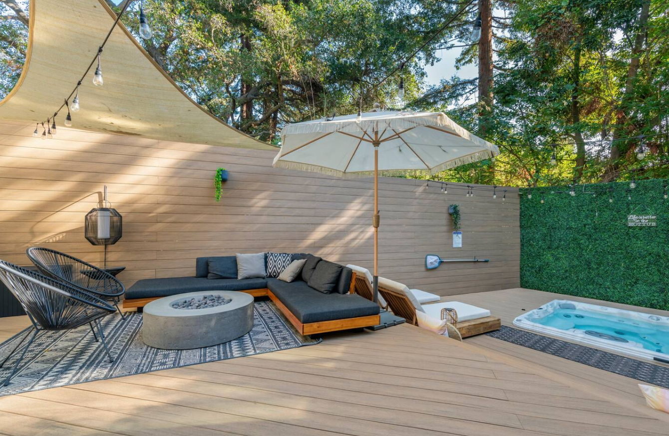 Outdoor patio with a sectional sofa, coffee table, lounge chairs, umbrella, hot tub, wooden deck, and string lights surrounded by trees.