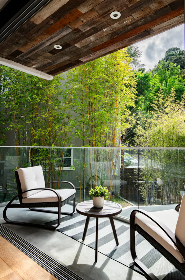 Modern balcony with glass railing, white cushioned chairs, a small round table with a potted plant, and lush green trees in the background under a wooden ceiling.
