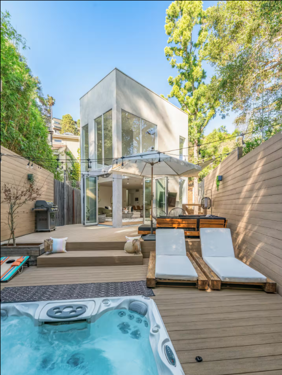 Modern backyard with hot tub, wooden deck, lounge chairs, umbrella, and a two-story white house surrounded by trees under a blue sky.