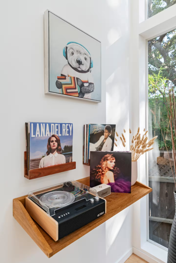 A vinyl record player on a wooden shelf with LP records, a white speaker, and a potted plant with dried grasses, with paintings and photographs on the wall and a large window in the background.
