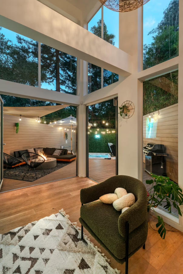 A cozy indoor living space with large windows and a view of an outdoor patio area with string lights, a hot tub, and a barbecue grill. Inside, there is a green armchair with plush pillows, a patterned rug, and a plant.