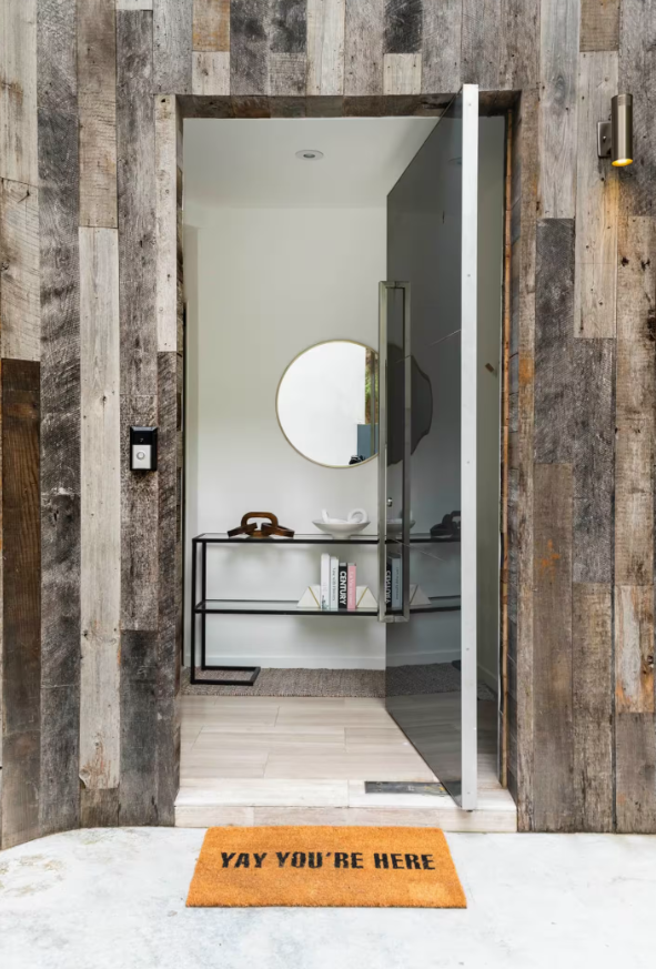 View through an open doorway into a modern entryway with white walls, a circular mirror, and a black console table with decorative items. Outside the door, there is an orange doormat that says "YAY YOU'RE HERE" on a concrete floor.