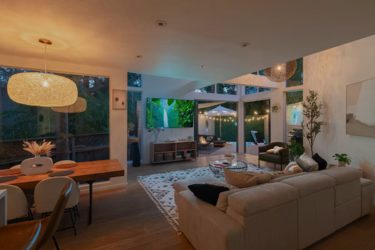 Modern living room and dining area with large windows, cozy seating, and string lights at dusk.