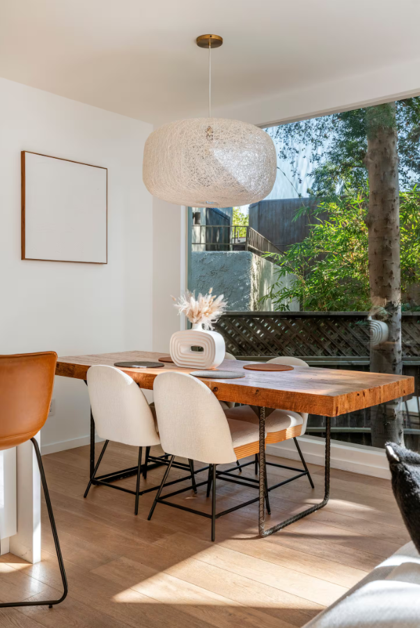 A dining room with a wooden table, three beige upholstered chairs, a brown chair, a white vase with dried flowers, a large white pendant light, and a window showing outdoor greenery.