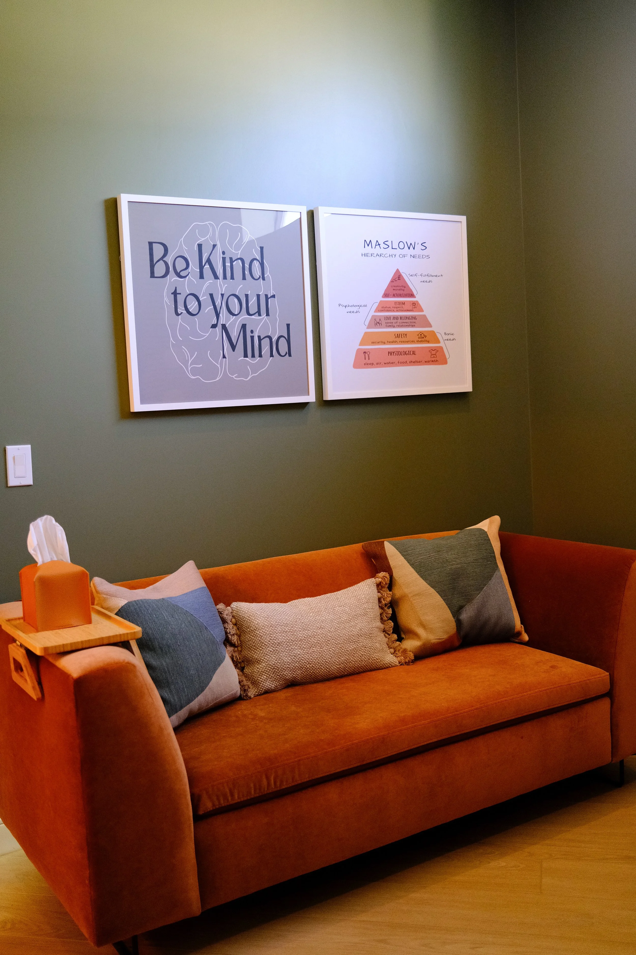 A cozy waiting area with an orange sofa, multiple pillows, a wooden side table with tissue box and hand sanitizer, green wall with two framed posters, one with the text "Be Kind to your Mind" and the other with Maslow's hierarchy of needs.
