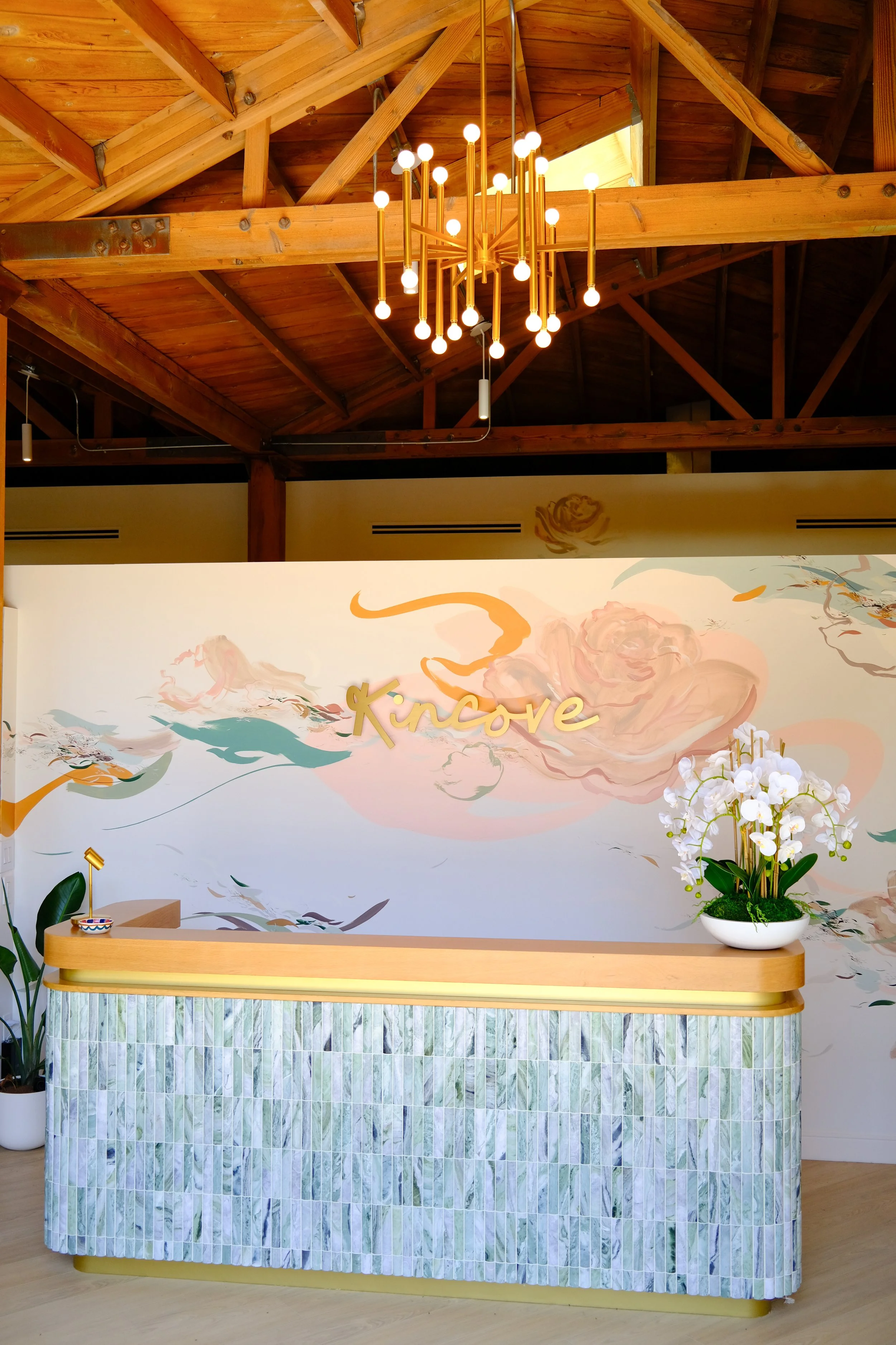 Interior of a reception area with a modern chandelier hanging from a wooden ceiling. The wall behind the reception desk features a floral mural with the word "Kinase" in gold lettering. The desk has a tiled front with pastel colors and a white orchid plant on the right side.