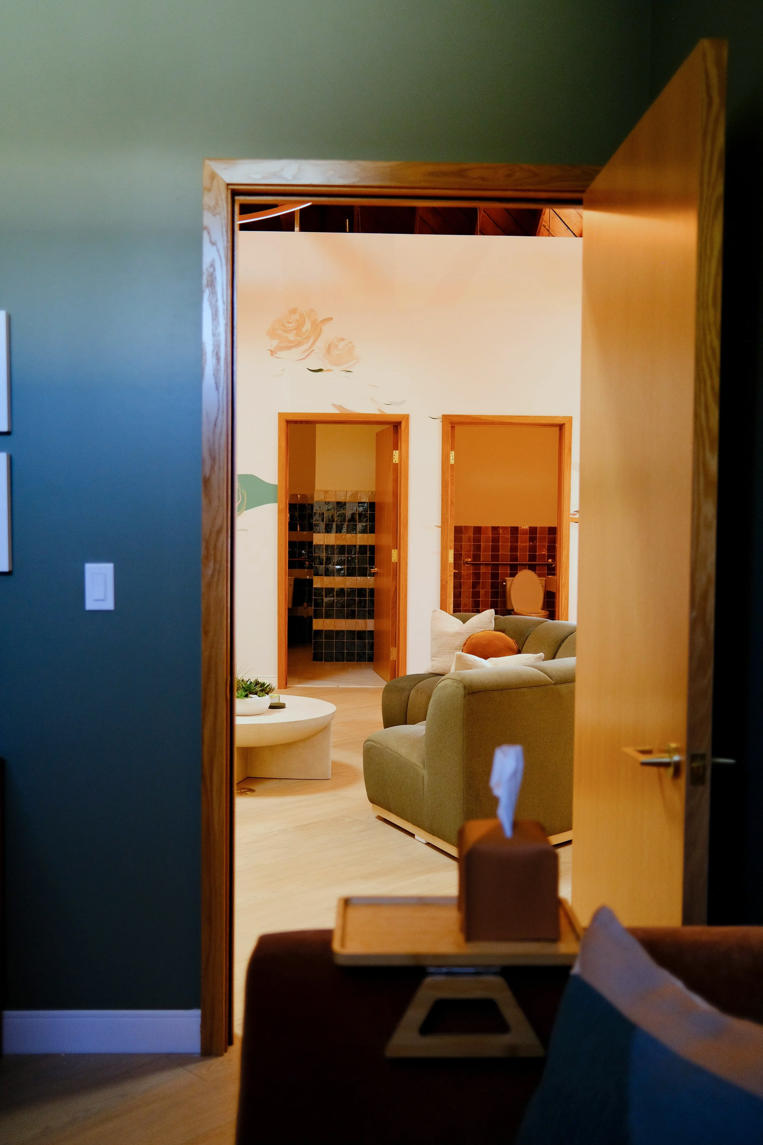 View through an open wooden door into a living room with a mustard sofa, a round white coffee table, and a hallway with tiled walls and a toilet.