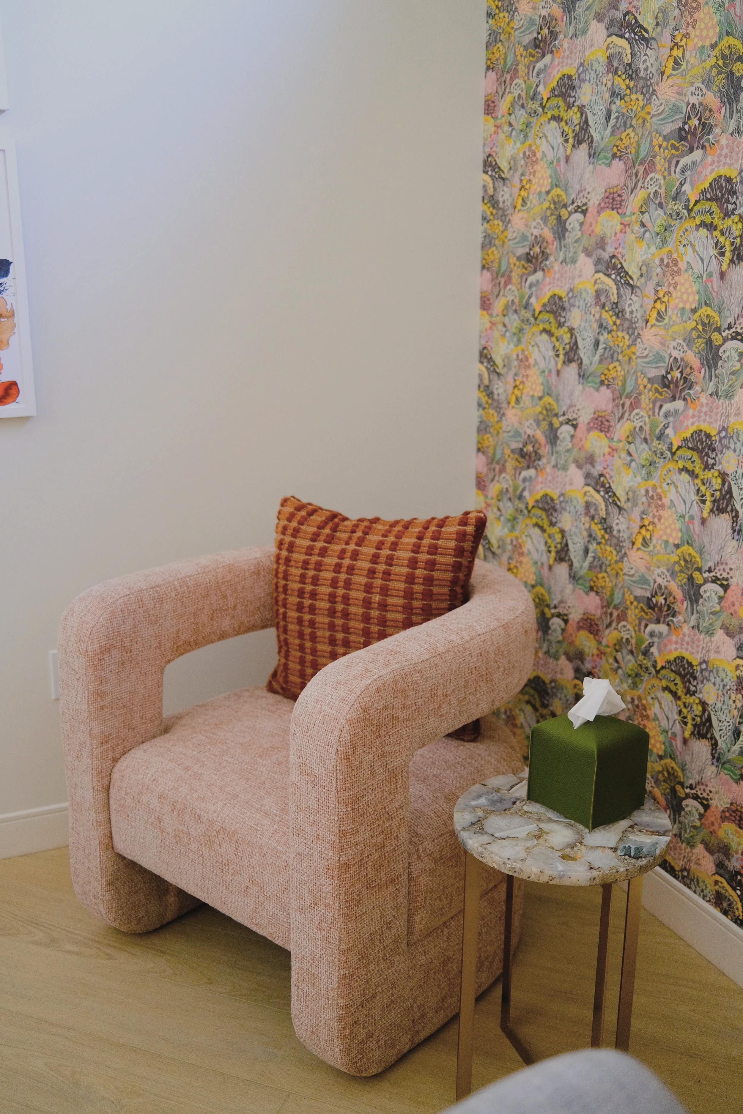 A pink upholstered armchair with a red and brown checked cushion against a white wall and a floral wallpapered accent wall, next to a small marble-topped side table with a green tissue box.
