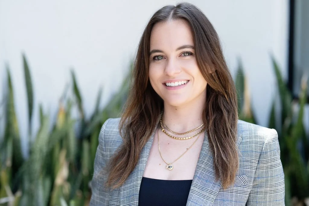 A woman with long brown hair, smiling, wearing a plaid blazer and layered necklaces, standing outdoors with green plants in the background.