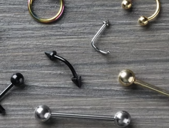 A smiling young man with tattoos, wearing a black apron and black gloves, poses with hands near his head against a plain background.