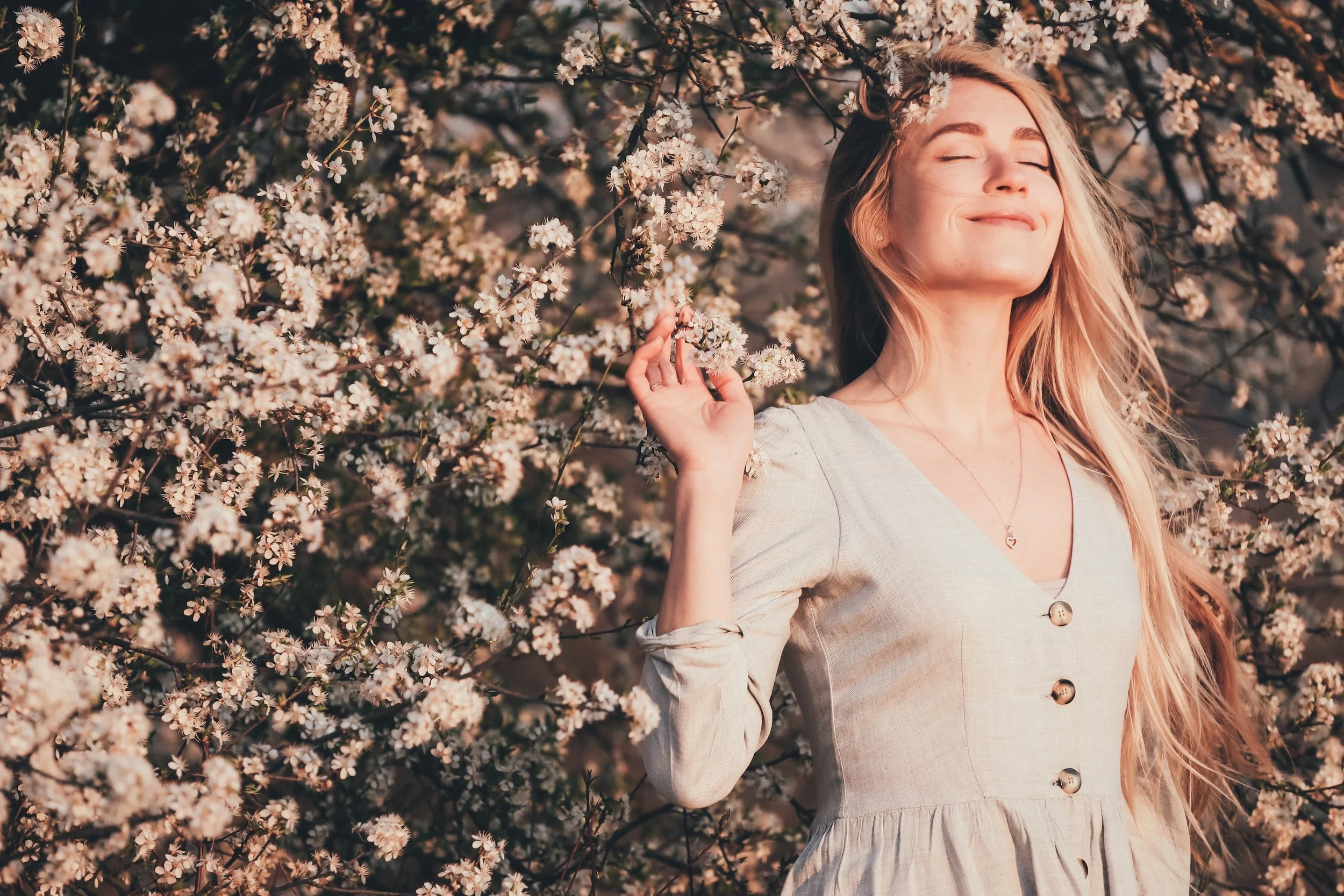 Woman with long hair enjoying cherry blossoms in sunlight, wearing a light dress, eyes closed in contentment.