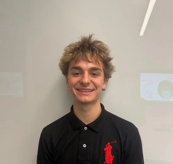 A young man with messy blond hair wearing a black polo shirt with a red logo, smiling, standing in front of a whiteboard or wall.