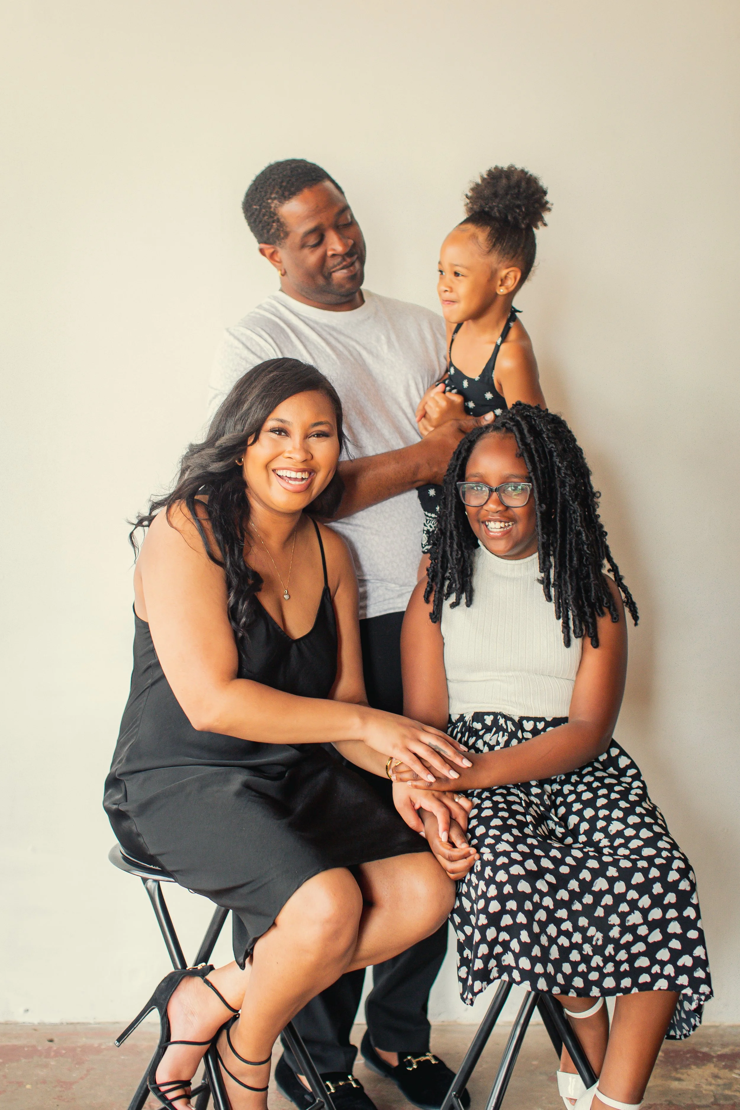 A happy family of four posing together indoors. A woman wearing a black dress and high heels, a girl with glasses and a patterned skirt, a man in a gray T-shirt, and another girl with a high curly ponytail in a black and white dress.