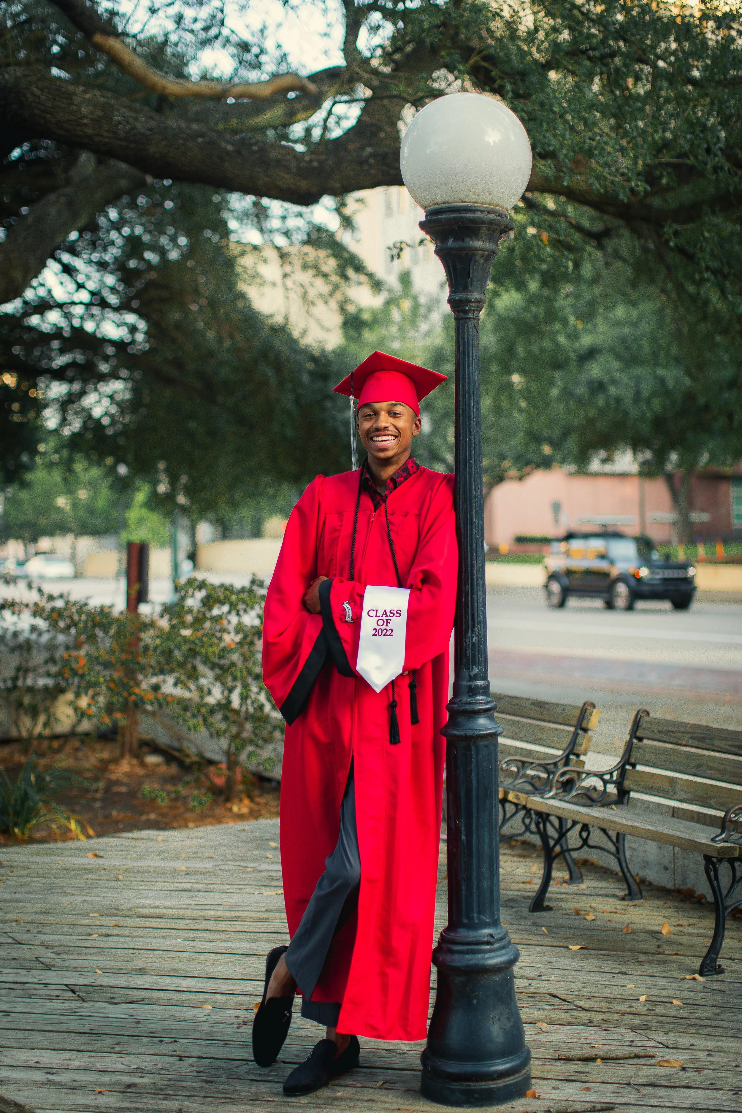 A young man in a red graduation gown and cap, smiling and holding a diploma, standing next to a lamppost on a park-like setting with trees, benches, and a street in the background.