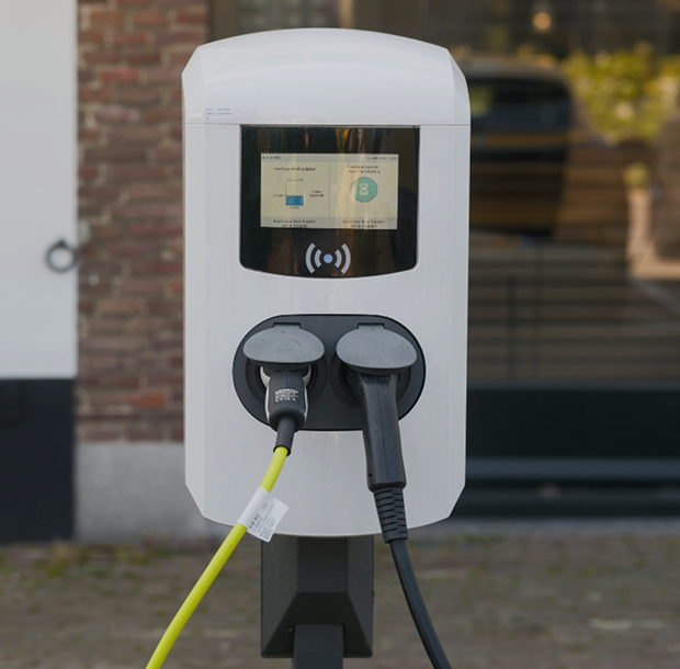 Elektrische laadpaal voor auto's met twee oplaadkabels, geplaatst buiten bij een gebouw.
