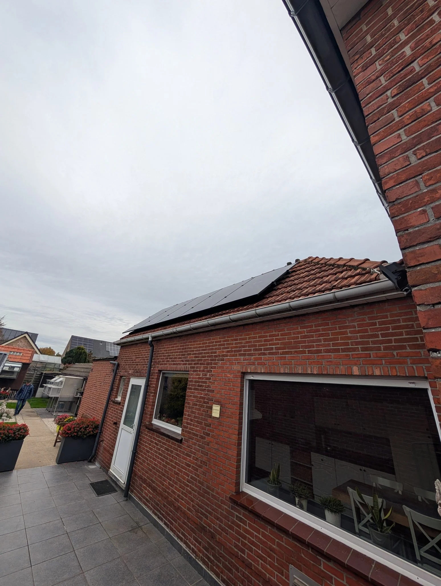 De gevel van een rood bakstenen huis met zonnepanelen op het dak en een tuin met planten en mensen op een pad.