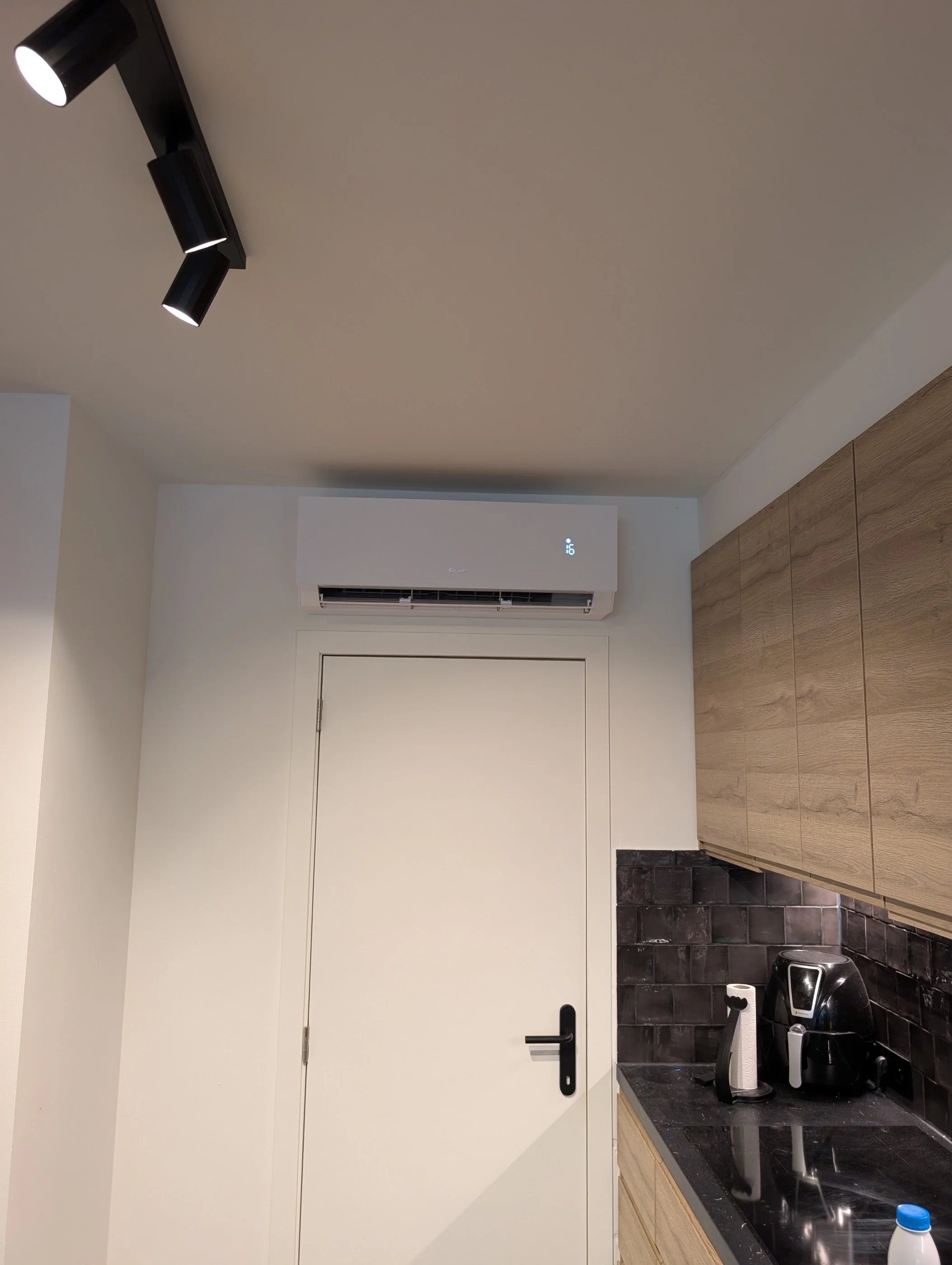 Keuken met een witte deur, houten kast, een koffiezetapparaat en een waterkoker op het zwartstenen werkblad, zwarte tegelachtergrond, plafond met drie zwarte spotlights.