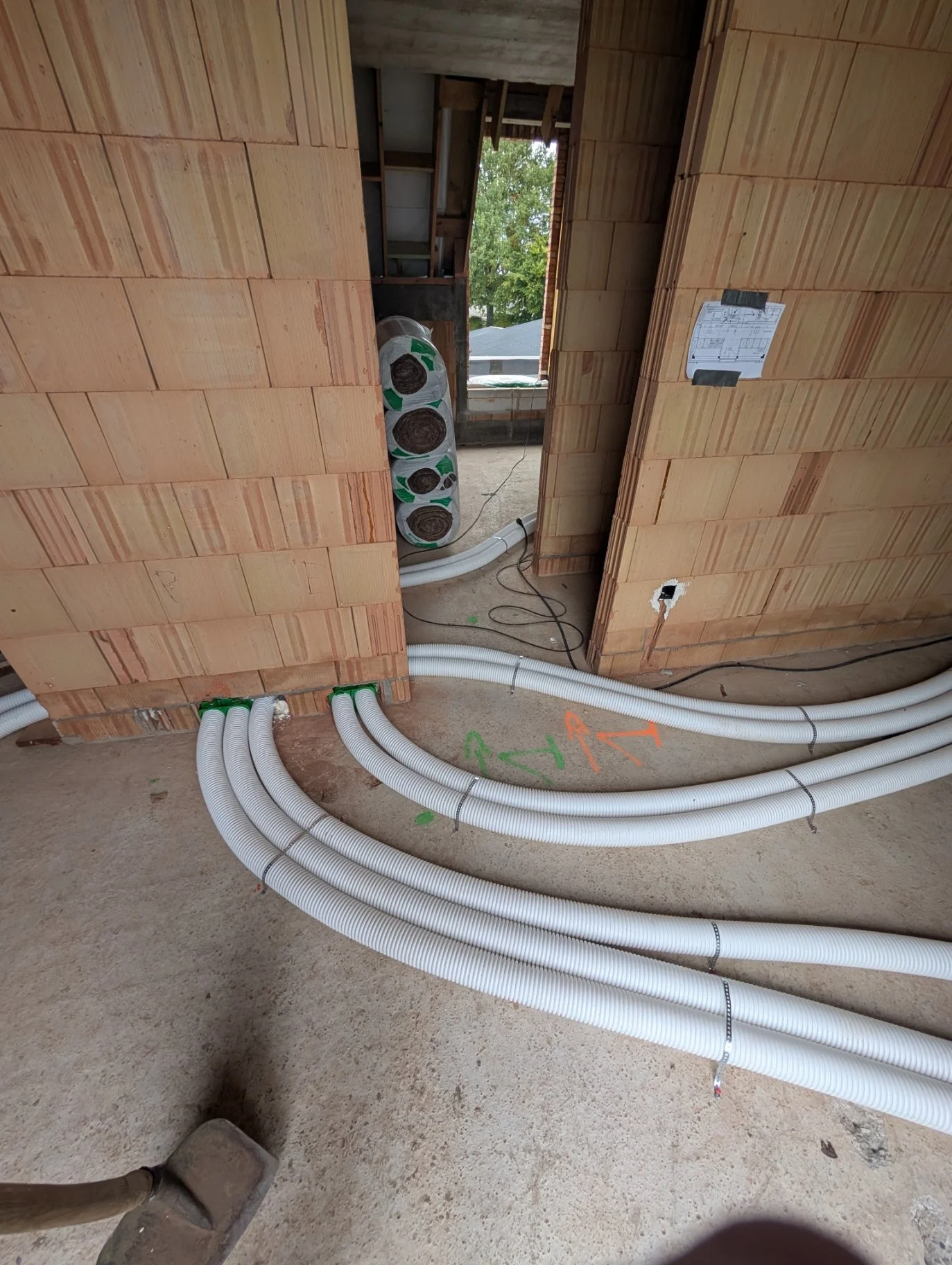 Binnen in een bouwplaats met zicht op geïnstalleerde witte flexibele leidingen op de vloer, en onafgewerkte muren van bakstenen. Een raam en nieuwe groene bomen zijn buiten zichtbaar.