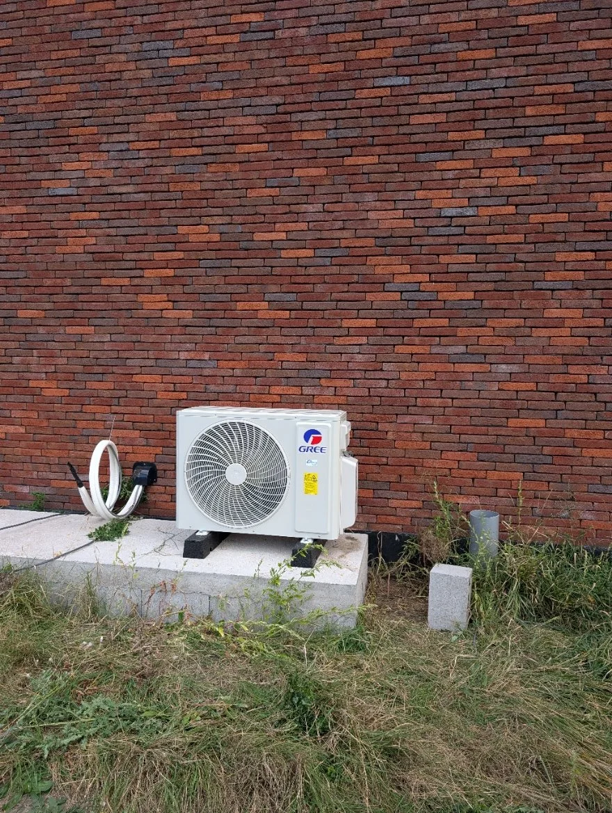 Een buiten unit van een airconditioningsysteem op een betonnen fundering naast een rode bakstenen muur, met een borduurigde slang en wat gras en stenen op de grond eromheen.