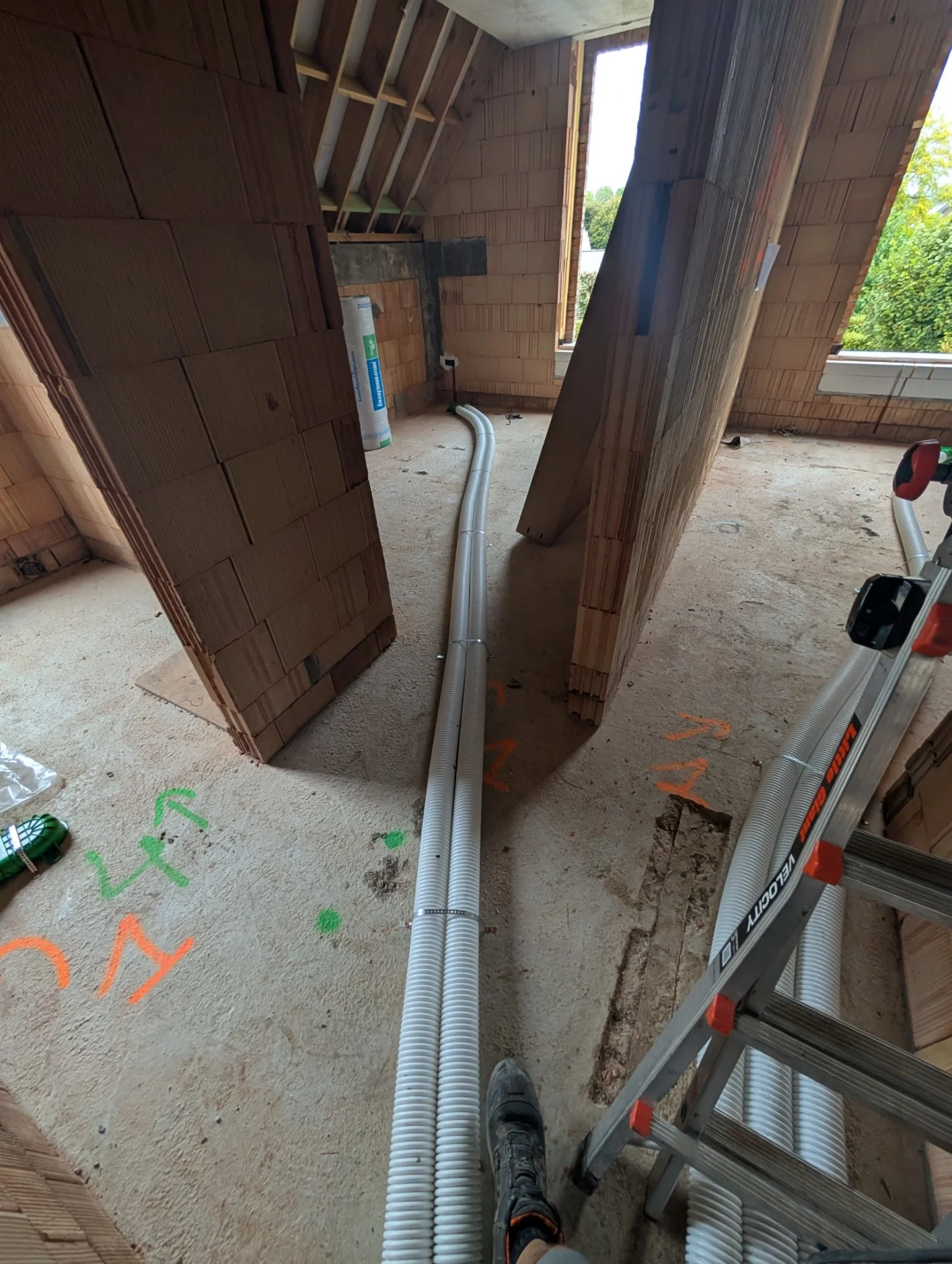 Interieur van een woning onder constructie, met geïnstalleerde ventilatie- of waterleidingen op de vloer, omgeven door onafgewerkte muren en een ladder.