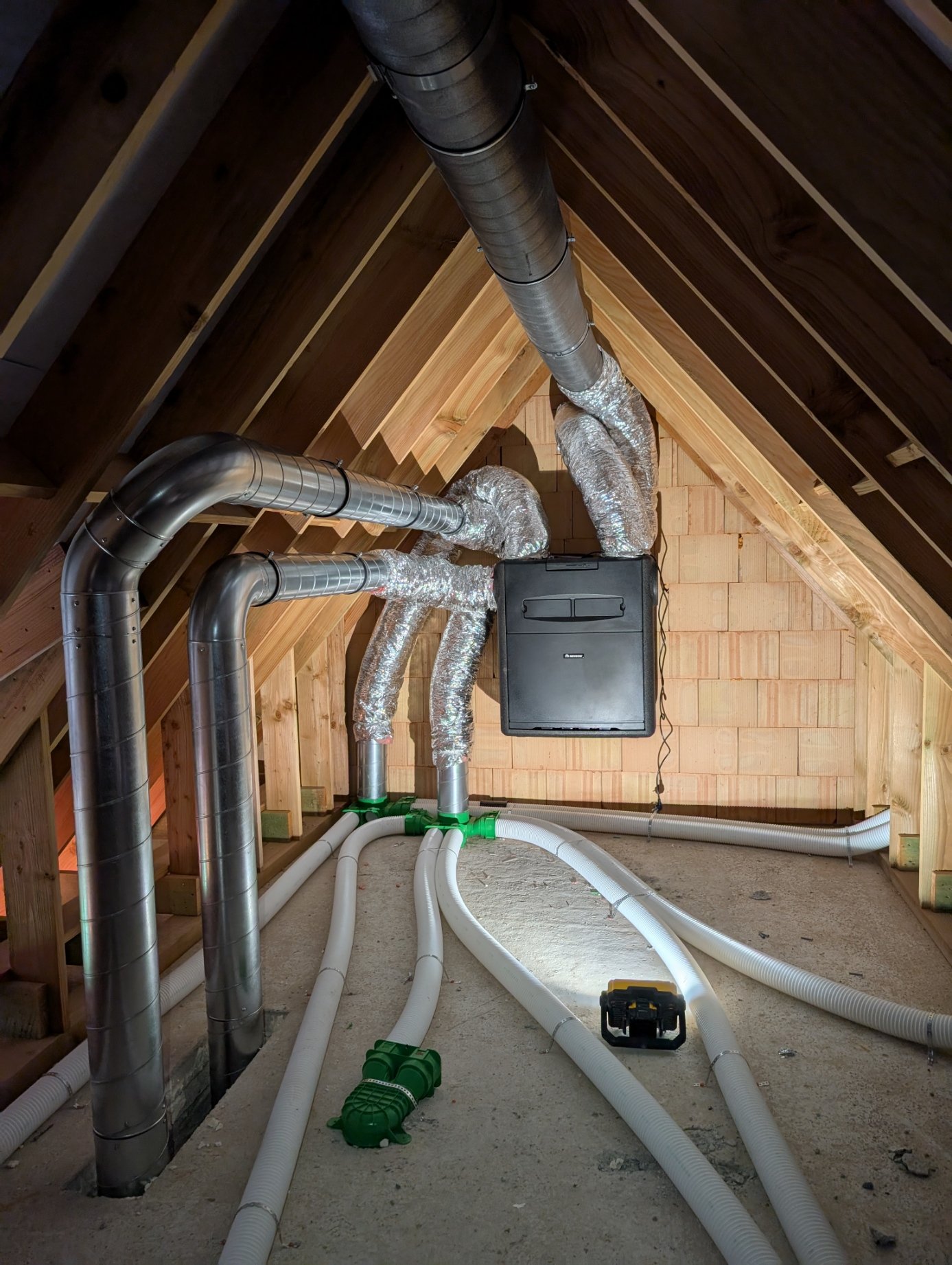 Installatie van ventilatie- en afzuigsysteem op zolder met metalen en plastic buizen, een ventilator en isolatie op de buizen.