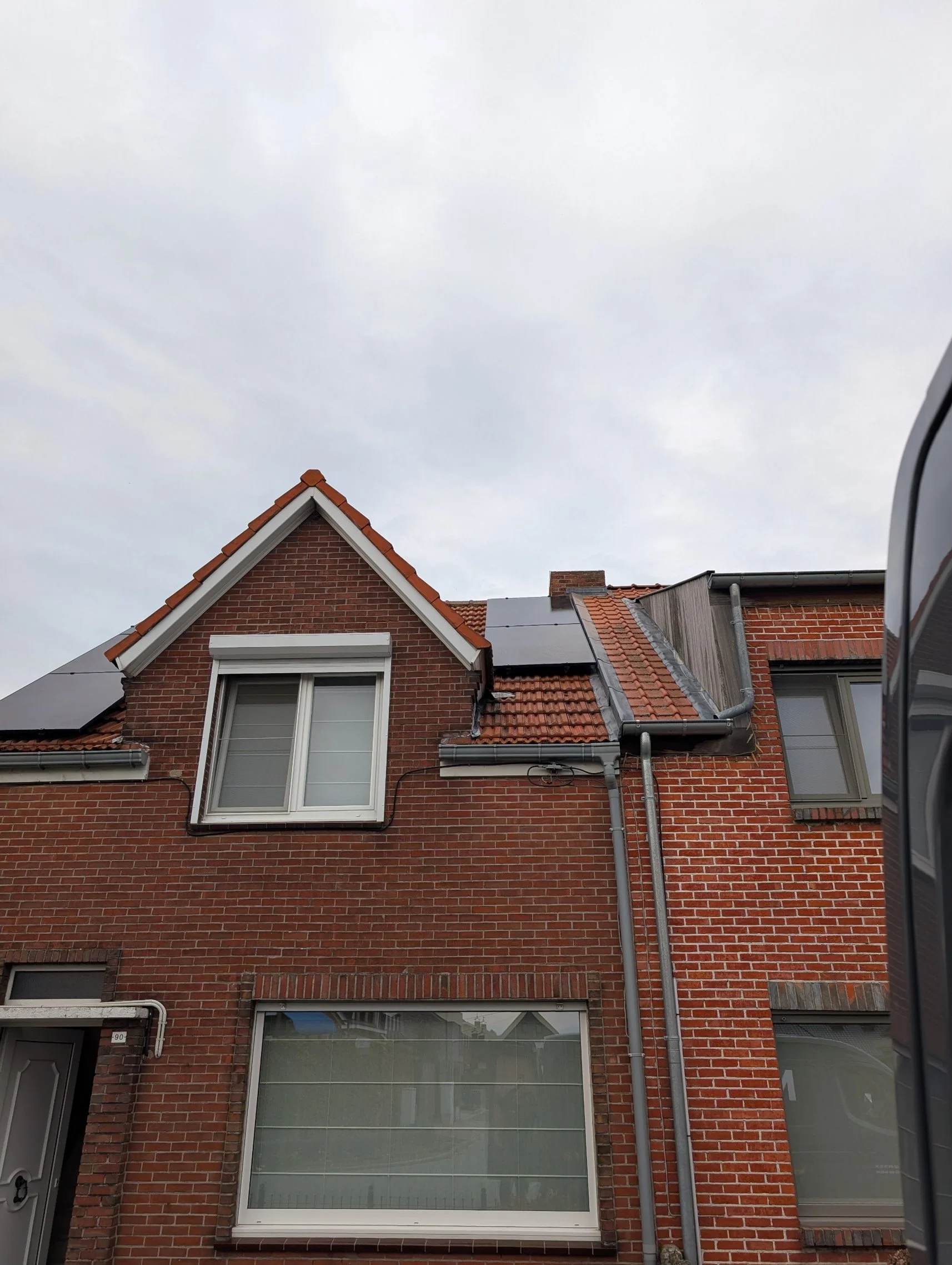 Een gevel van een rood bakstenen huis met dakkapellen en zonnepanelen. Een deel van een auto is aan de rechterkant zichtbaar.