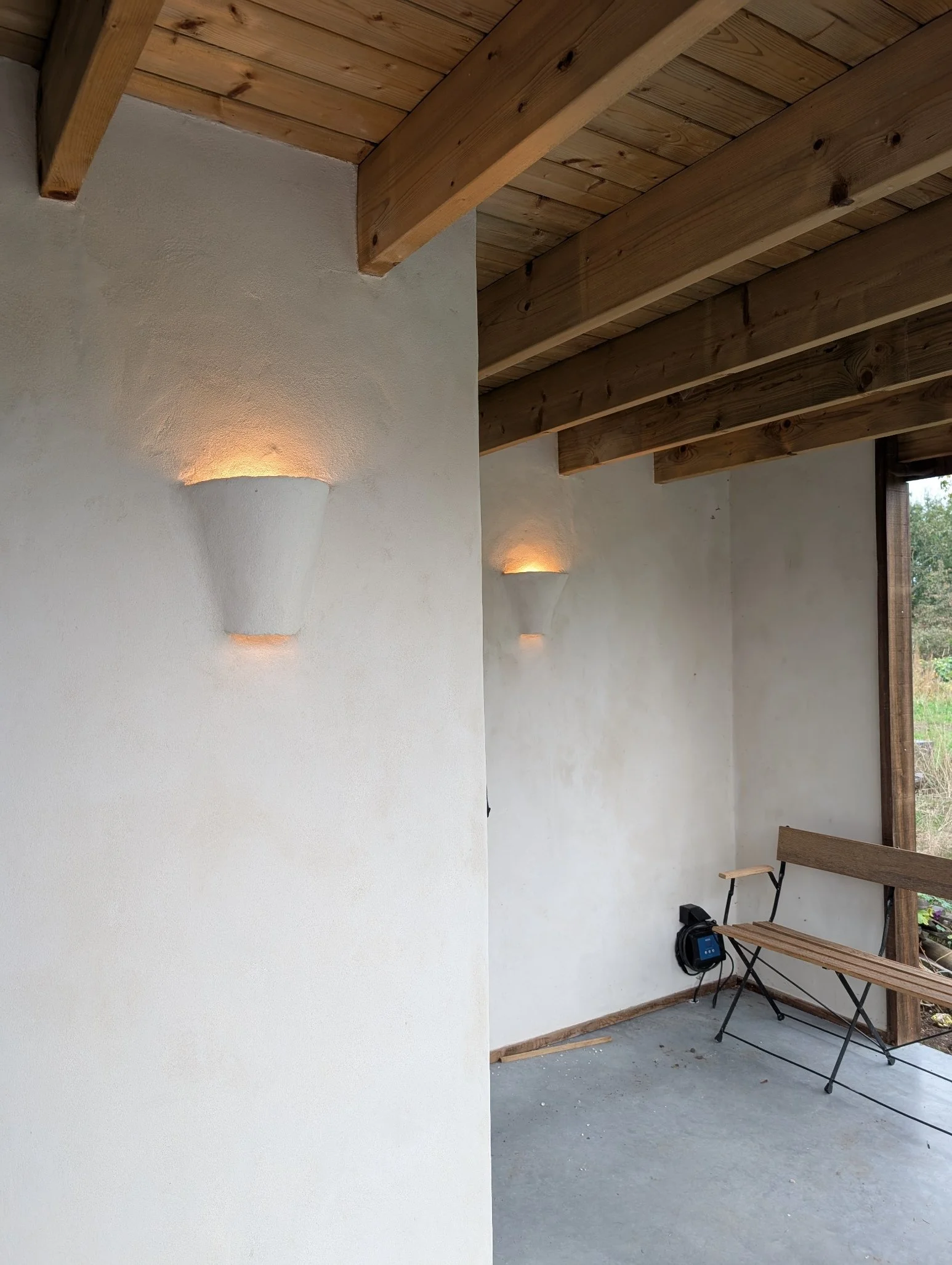 Interieur van een huis met witte betonnen muren, houten plafondbalken, en een kleine houten bank naast een raam.