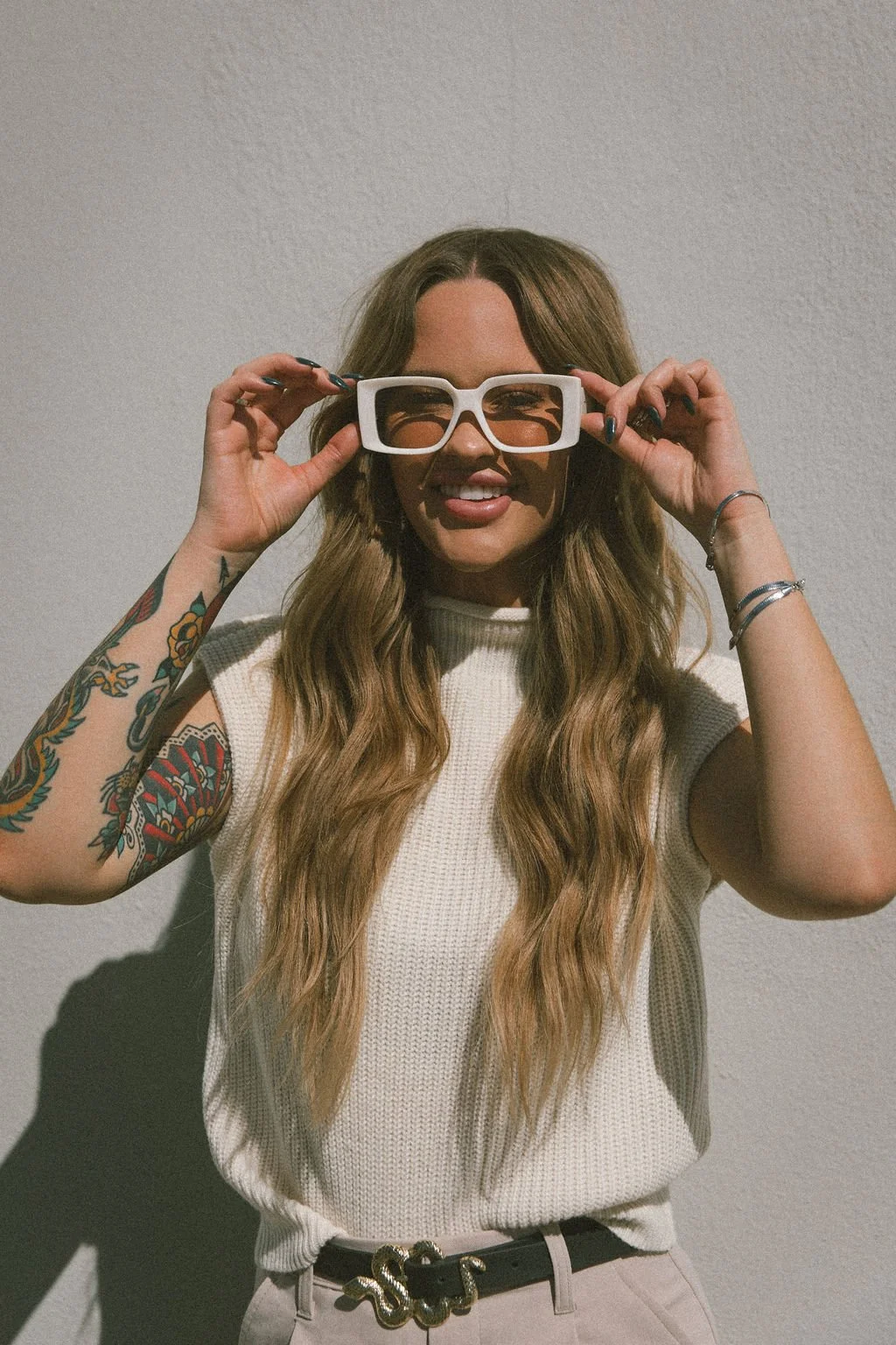 This image shows Emma smiling at the camera. She is wearing white retro sunglasses and is holding the frame with both of er hands. She is wearing a cream colored top and has long blonde hair.