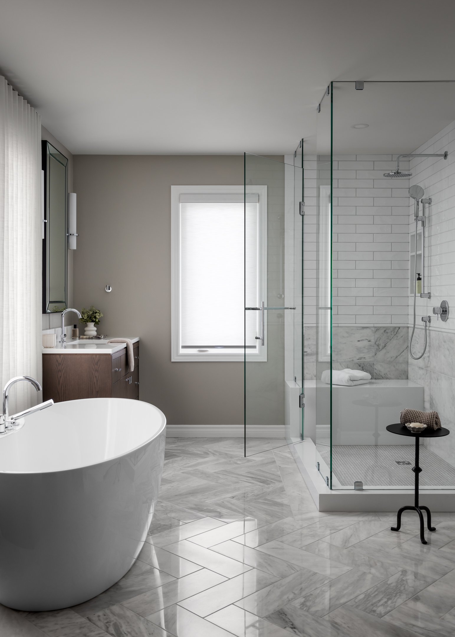 Elegant and timeless primary bathroom with a large soaker tub, herringbone marble floors, all glass shower enclosure, warm wood vanity by Studio Moffatt, London Ontario