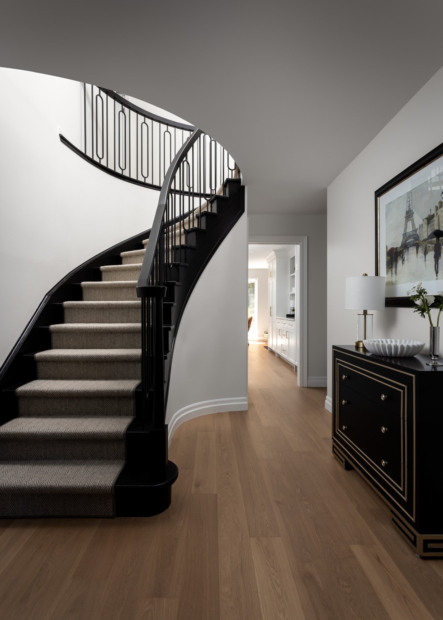 Residential renovation featuring a curved black painted staircase with herringbone carpet runner, metal balusters and wood railing.  White Oak hardwood flooring throughout the entrance foyer by Studio Moffatt in London, Ontario