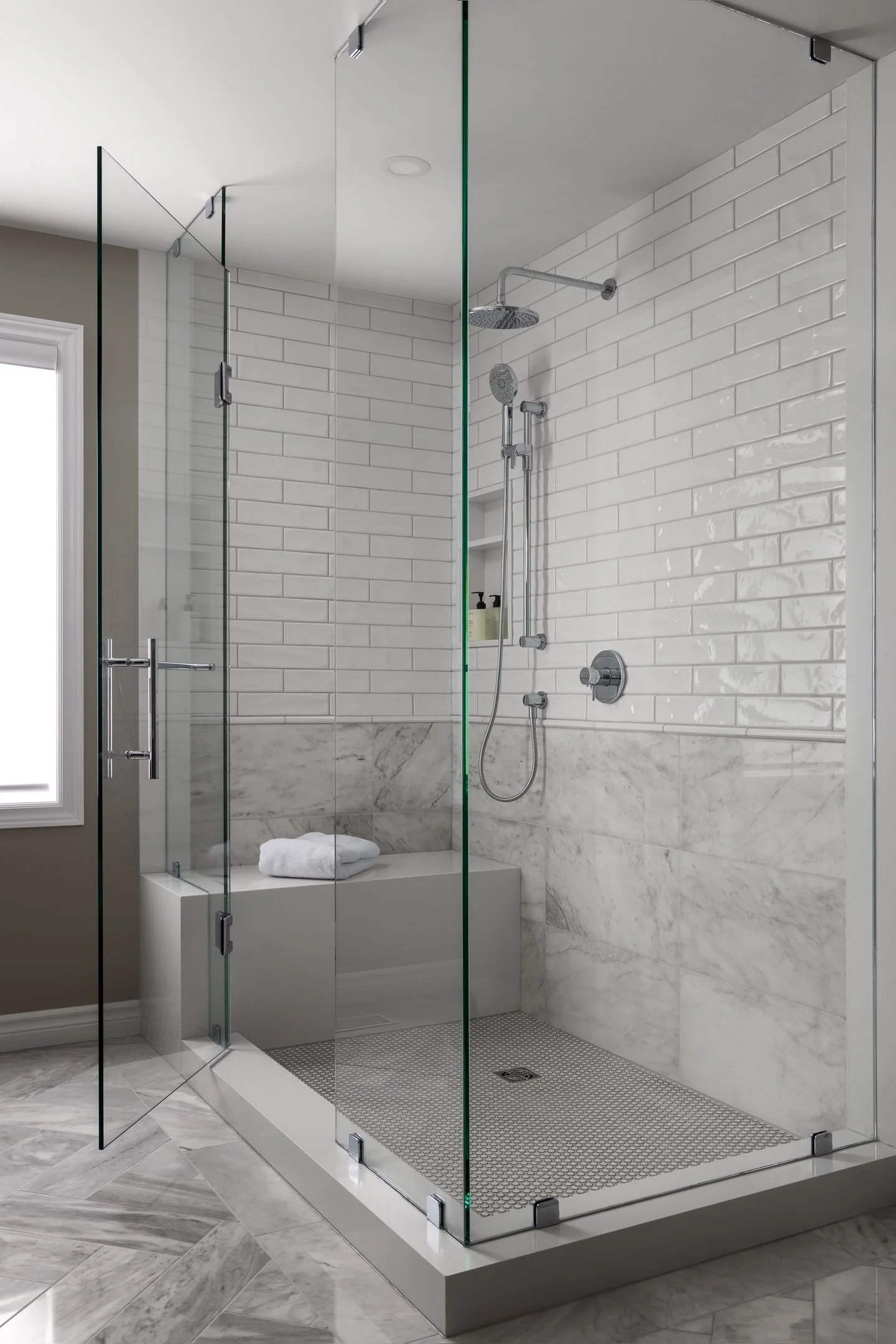 Timeless and elegant primary ensuite renovation featuring herringbone marble floors, glass shower enclosure with a combination of marble wall tile and a subway tile, and a white quartz shower bench by Studio Moffatt