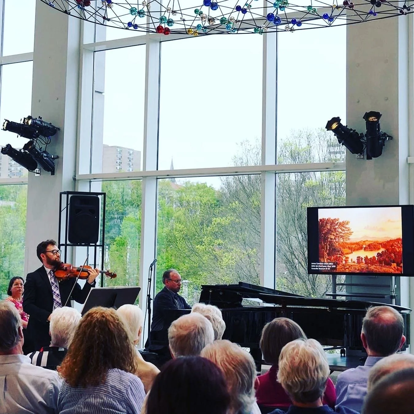 Violinist William Shaub performing in a large room with an audience, next to a grand piano. The room has large windows with a view of trees outside. A screen displays a landscape painting. The ceiling has a decorative structure with colored elements.