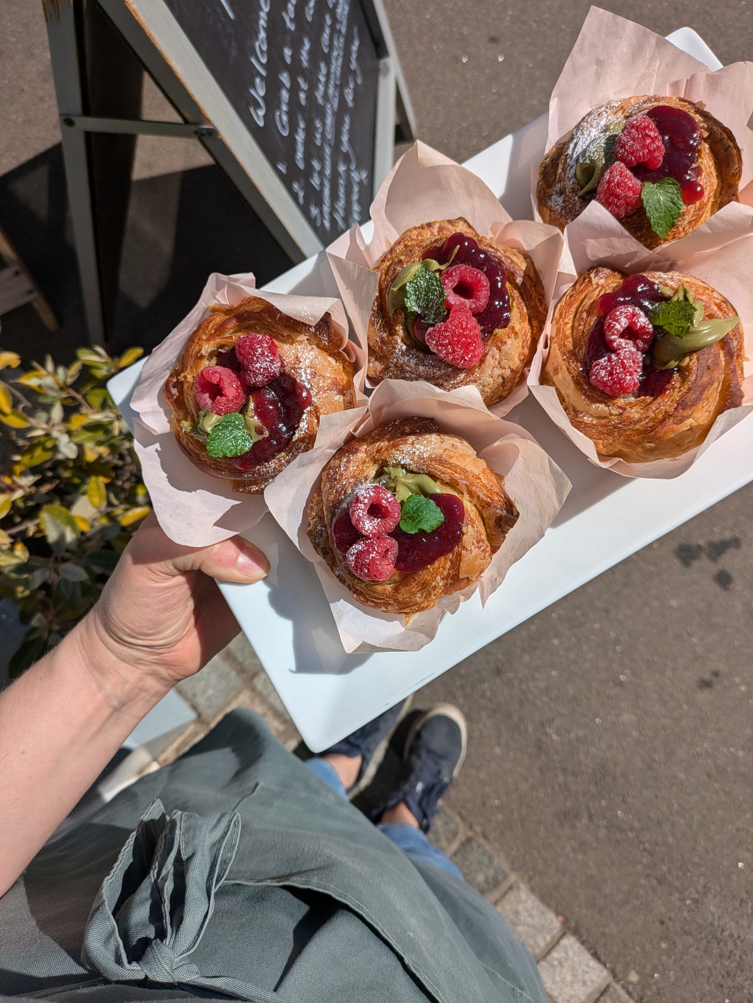 Person holding a white tray with five pastries topped with raspberries, green leaves, and berry sauces outside on a sunny day.