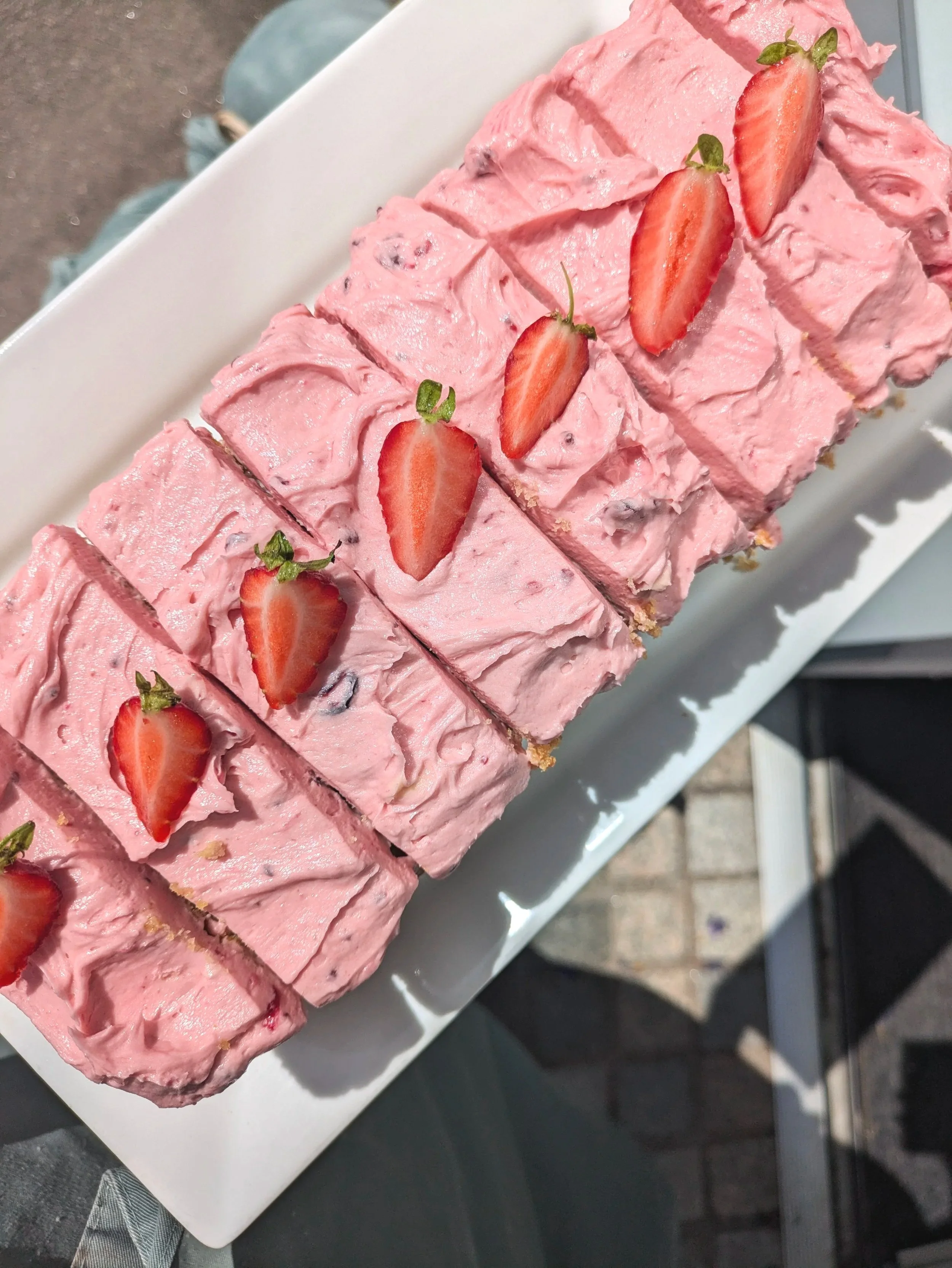 Slices of pink strawberry-flavored dessert topped with halved strawberries on a white rectangular plate.  Loaf cakes are baked and decorated in-house.