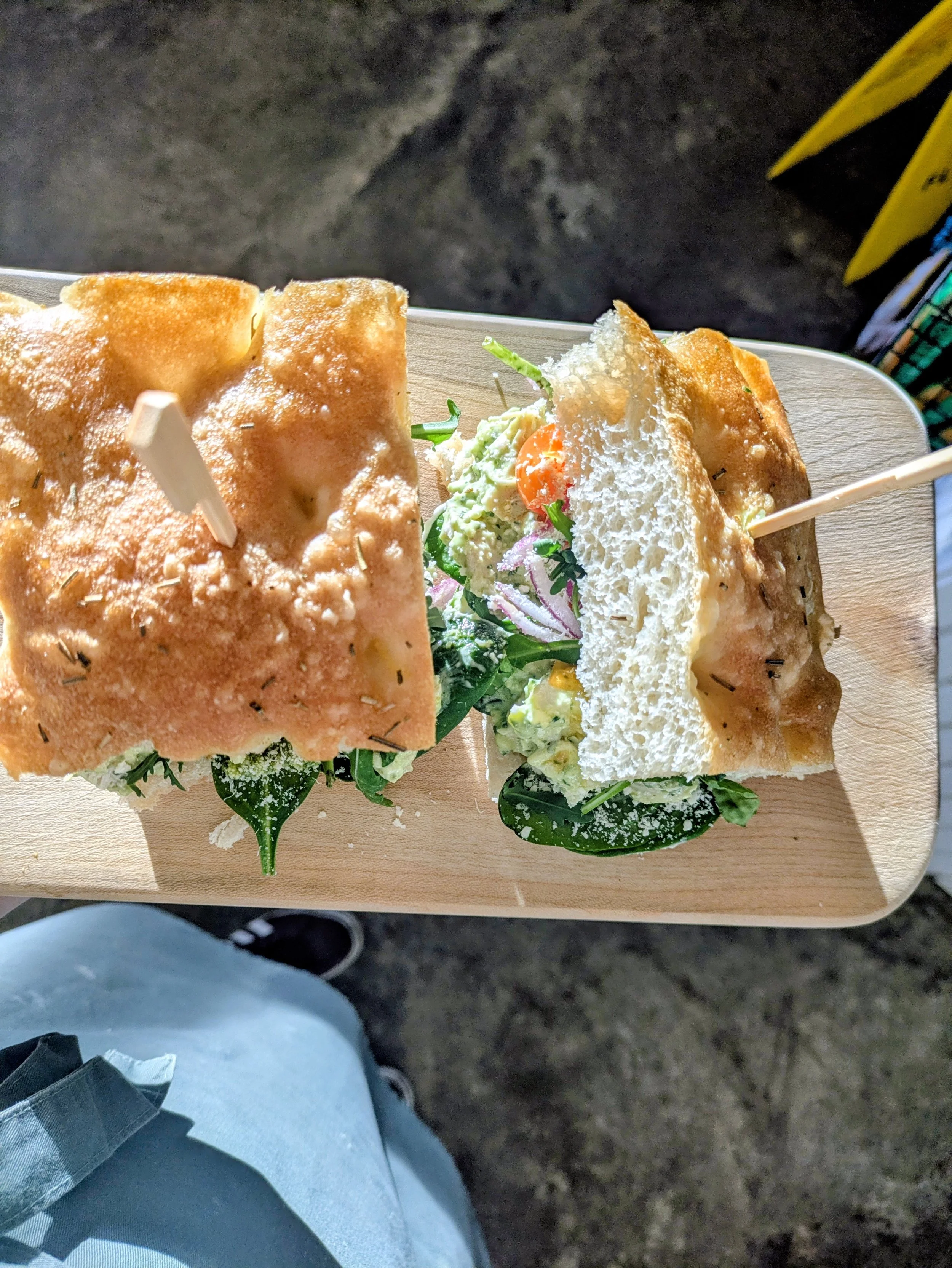 A sandwich with bread, vegetables, and greens on a wooden serving board.  Deli-style sandwiches prepared fresh and in small batches.